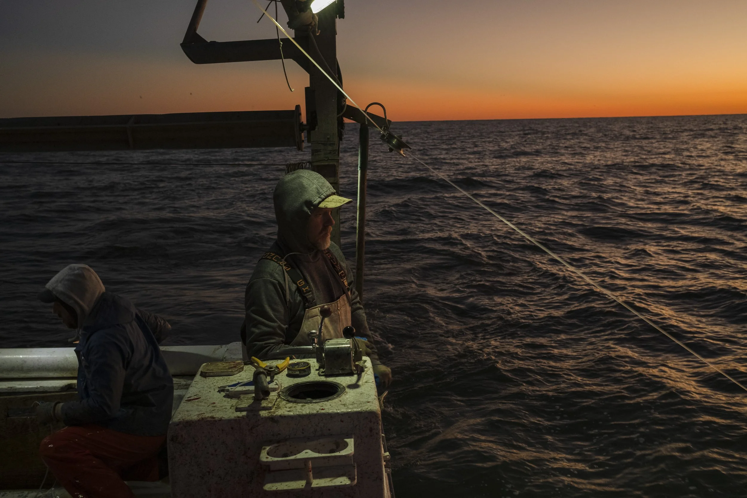 Two men on a boat fishing at sunset in the ocean, with one man standing and the other sitting, near fishing equipment.