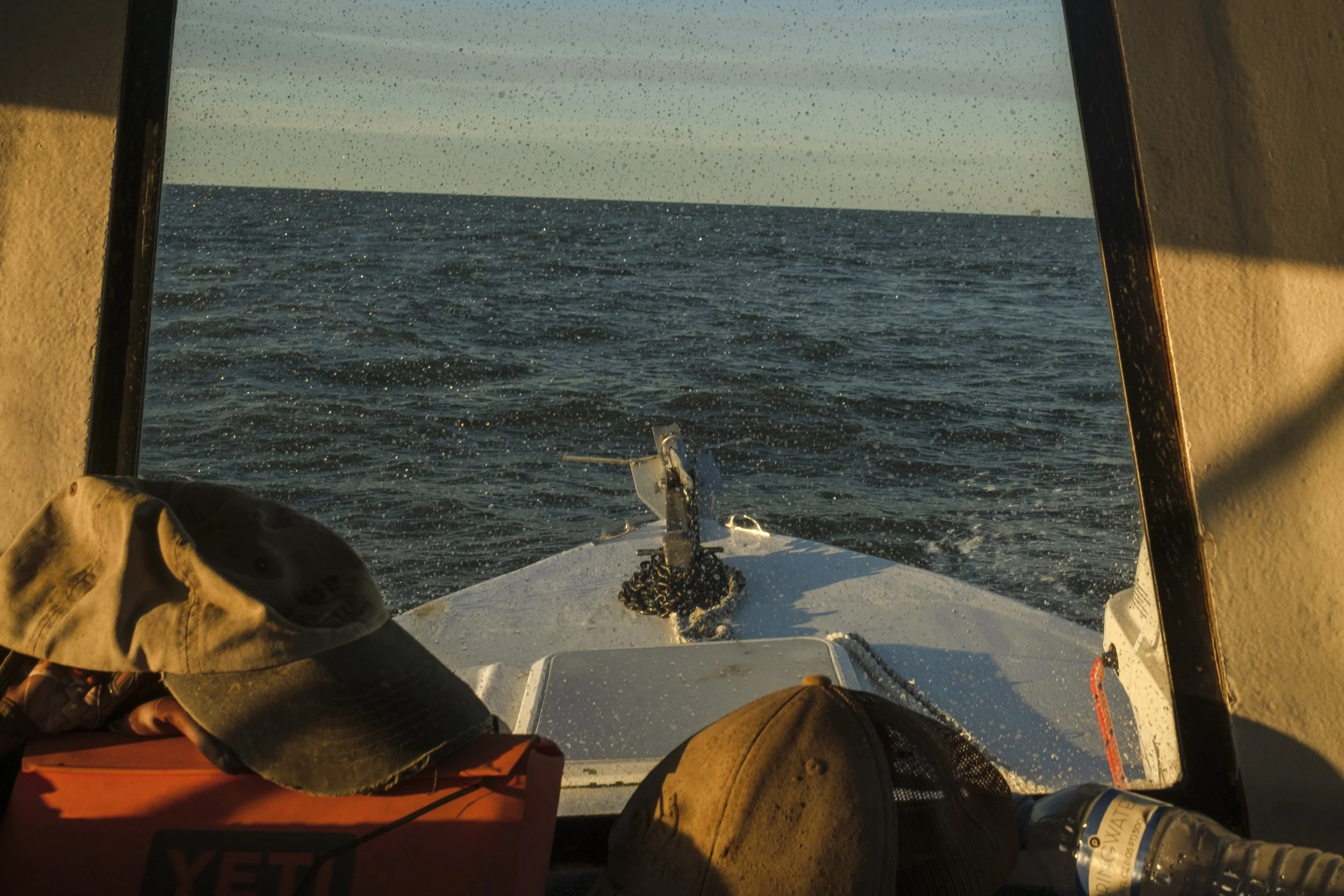 View from inside a boat looking out over the open ocean, with the boat's bow visible. The ocean is calm and expansive, with sunlight reflecting off the water. Inside the boat's cabin, there are various items including a cap, a water bottle, and a YET