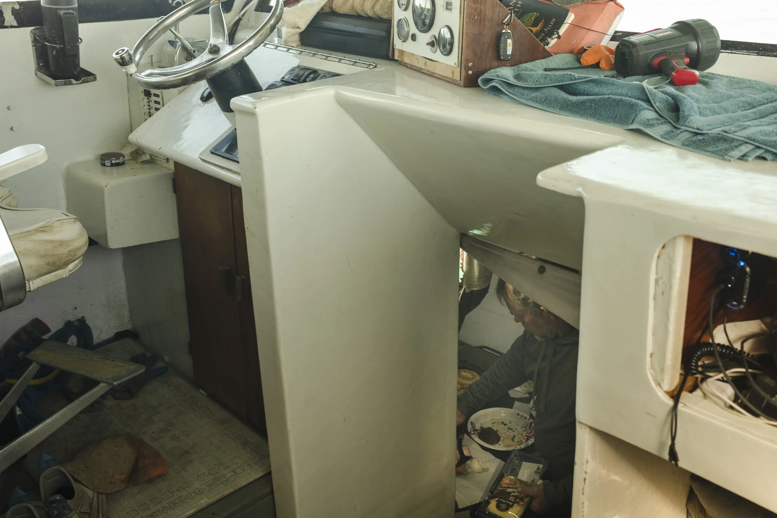 A person working under a boat cockpit or control panel, which has a beige surface with various gauges and tools. The person is partially visible beneath the panel, wearing glasses and gray clothing, holding a pizza box. The area appears cluttered wit