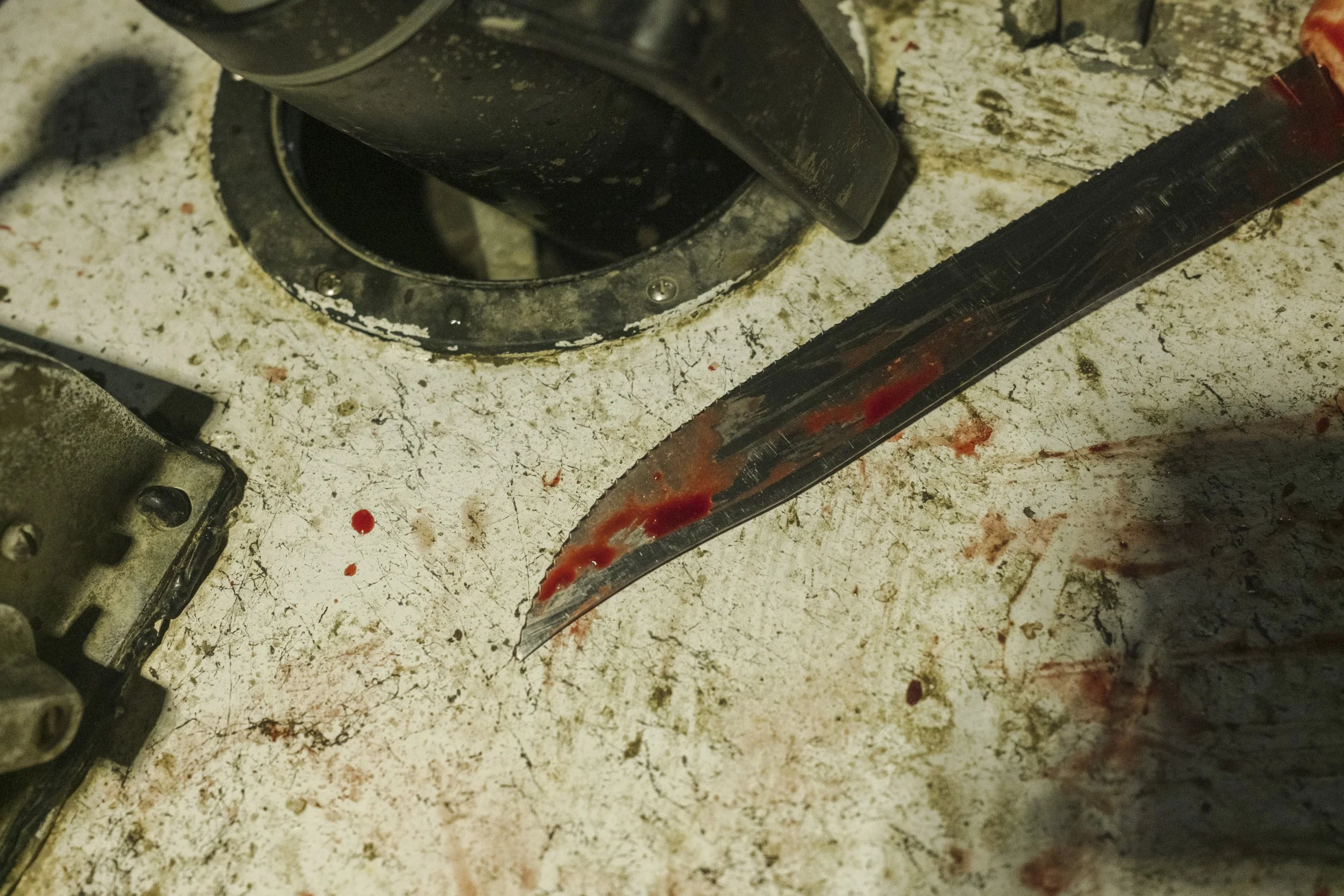 A dirty work table with bloodstains, a hacksaw with blood on the blade, and a machine part.