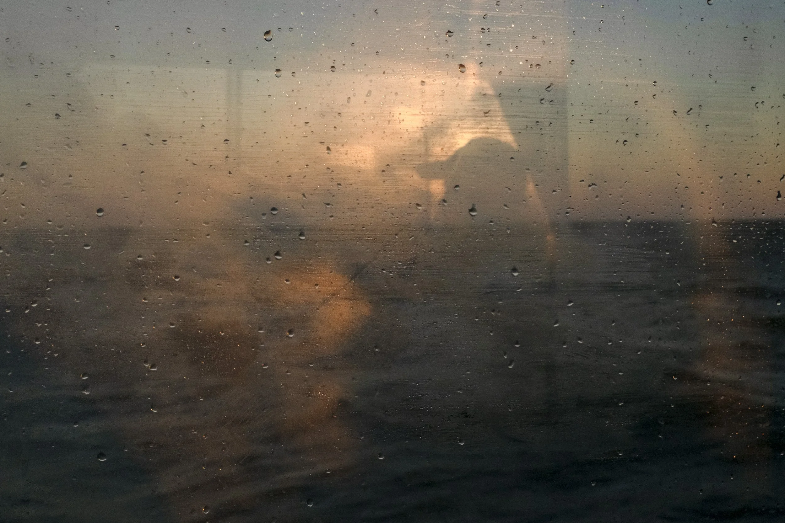 A view through a rain-covered window showing a sunset over the ocean with the silhouette of a person taking a photo.