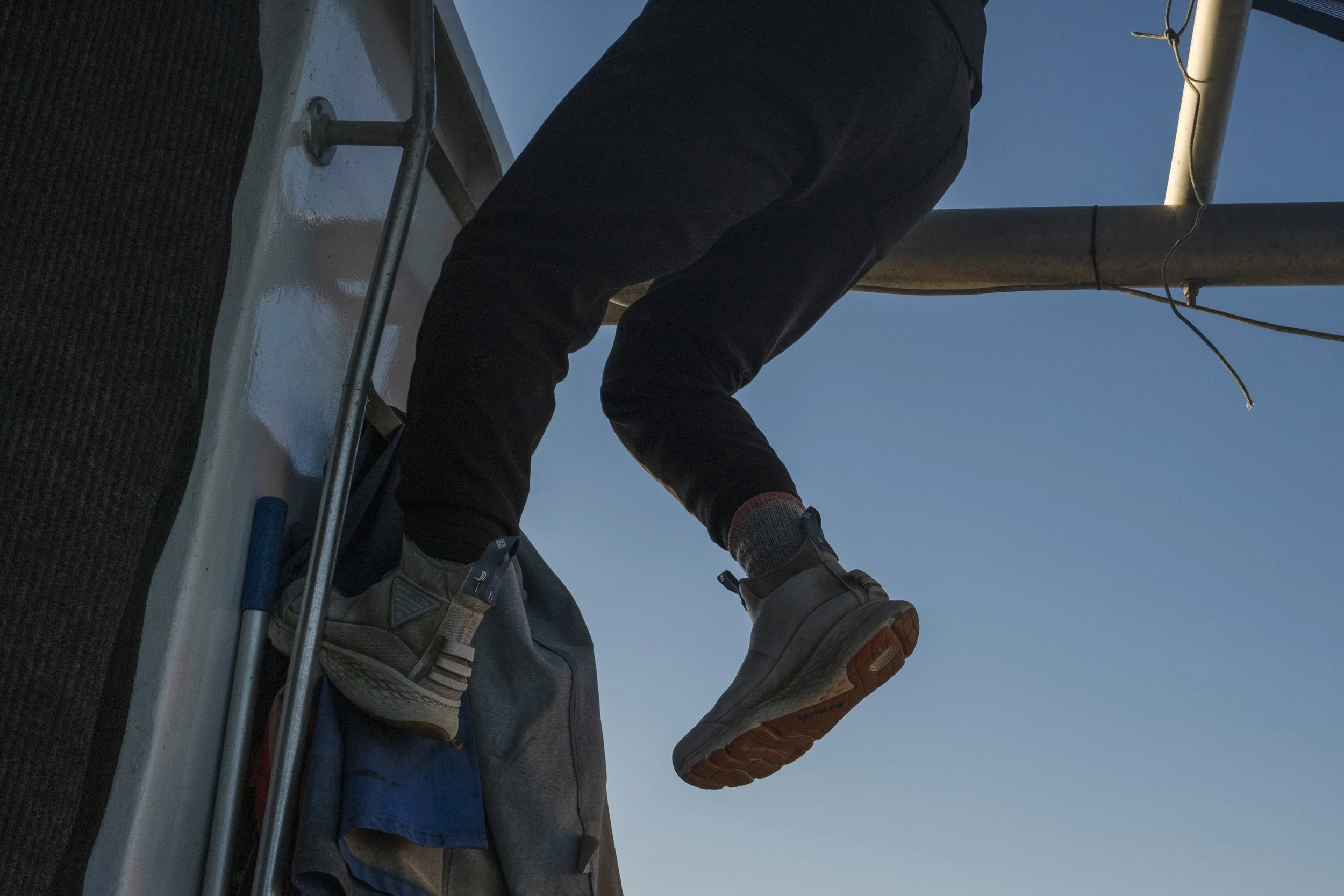 Close-up of a person climbing a structure, showing their legs and feet in sneakers, with a backpack and tools nearby, against a clear blue sky.