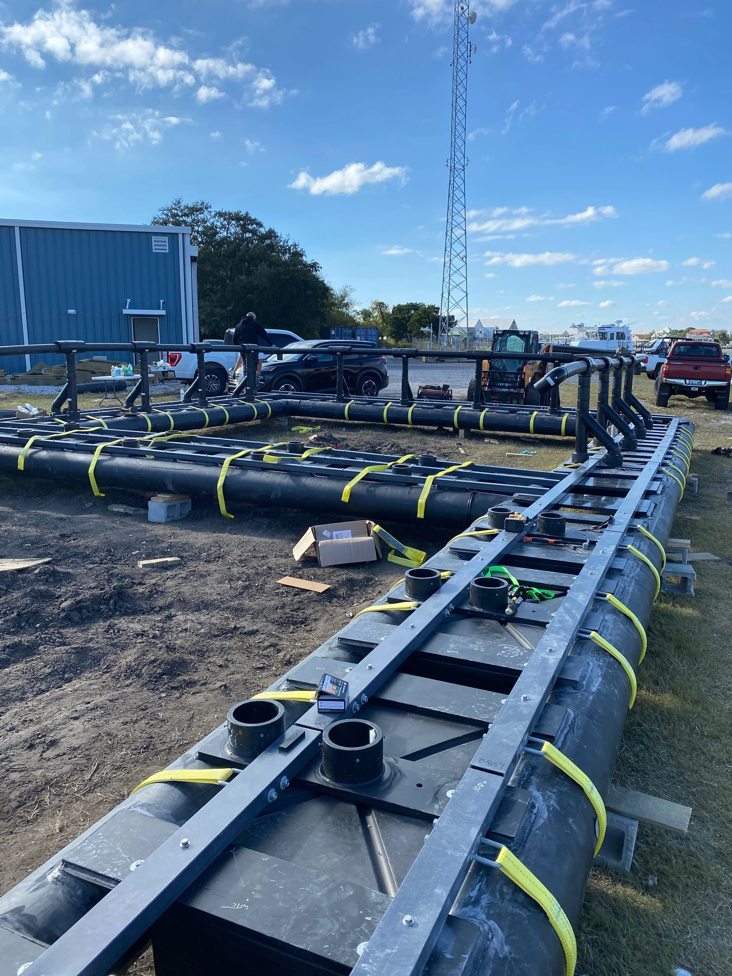 Construction site with large black pipe framework being installed, yellow straps securing the pipes, a man working near parked cars, a blue building, a telecommunications tower, and a partly cloudy sky.