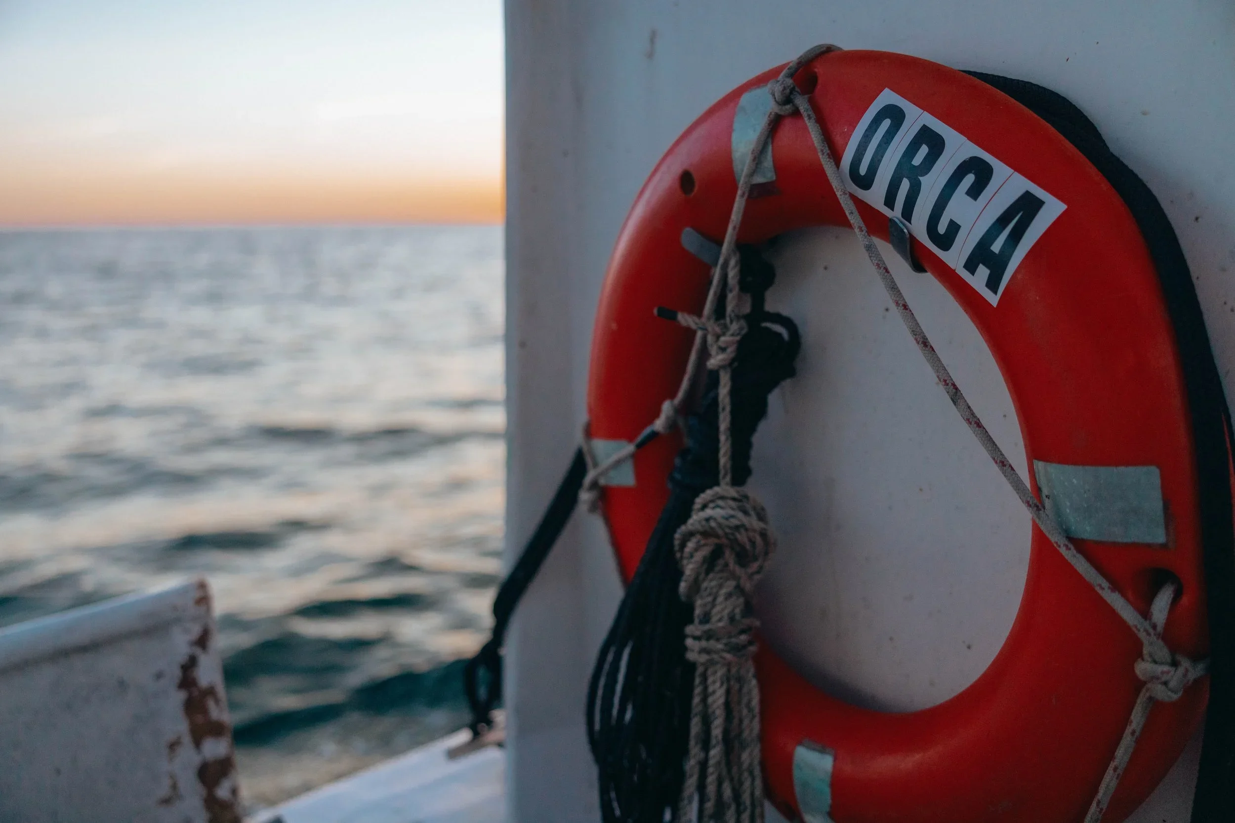 Close-up of an orange life ring with the word 'URCA' attached to the side of a boat, with the ocean and a sunset or sunrise in the background.
