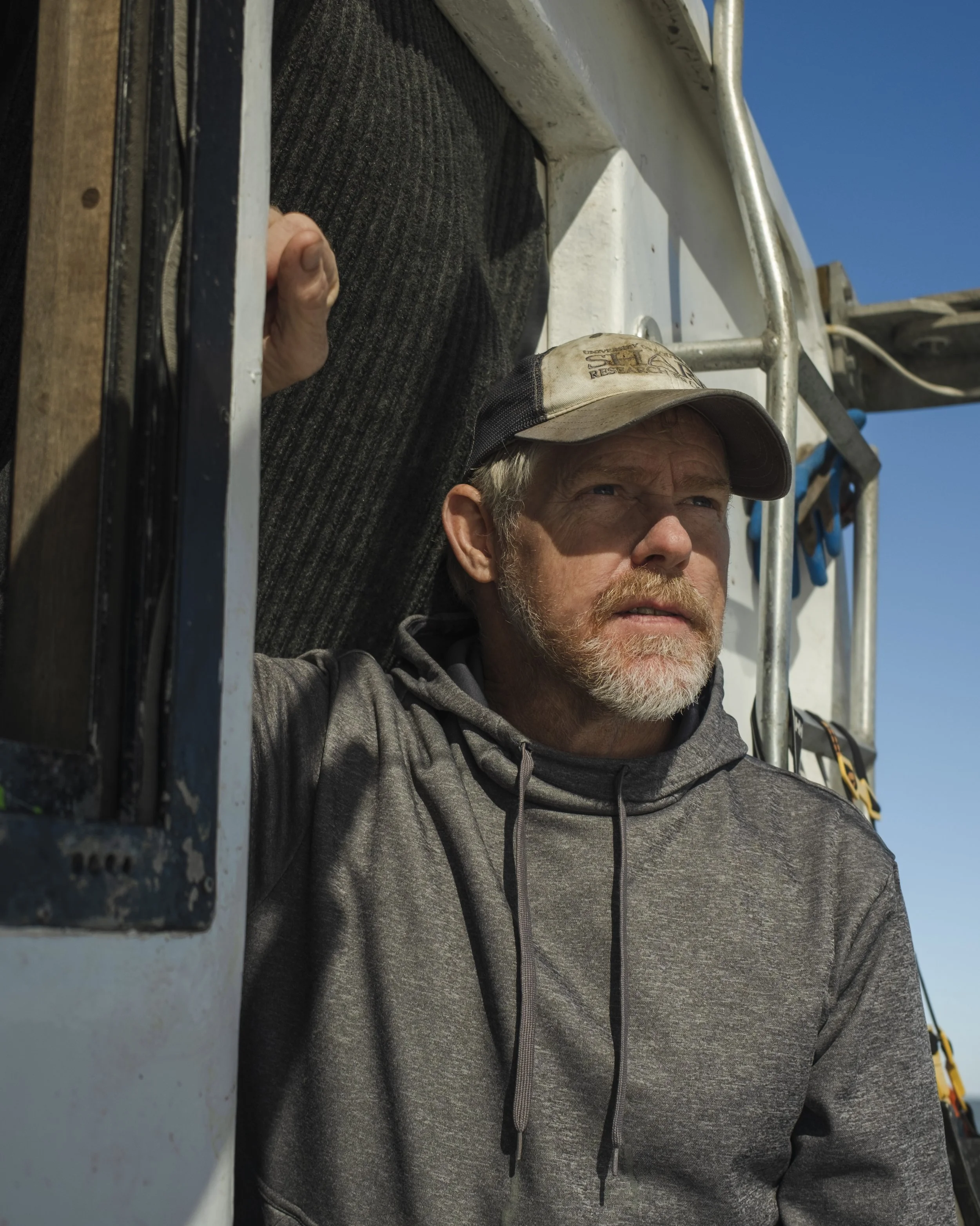 A man with a beard and mustache wearing a gray hoodie and a cap looks out from a window of a boat or vehicle against a clear blue sky.