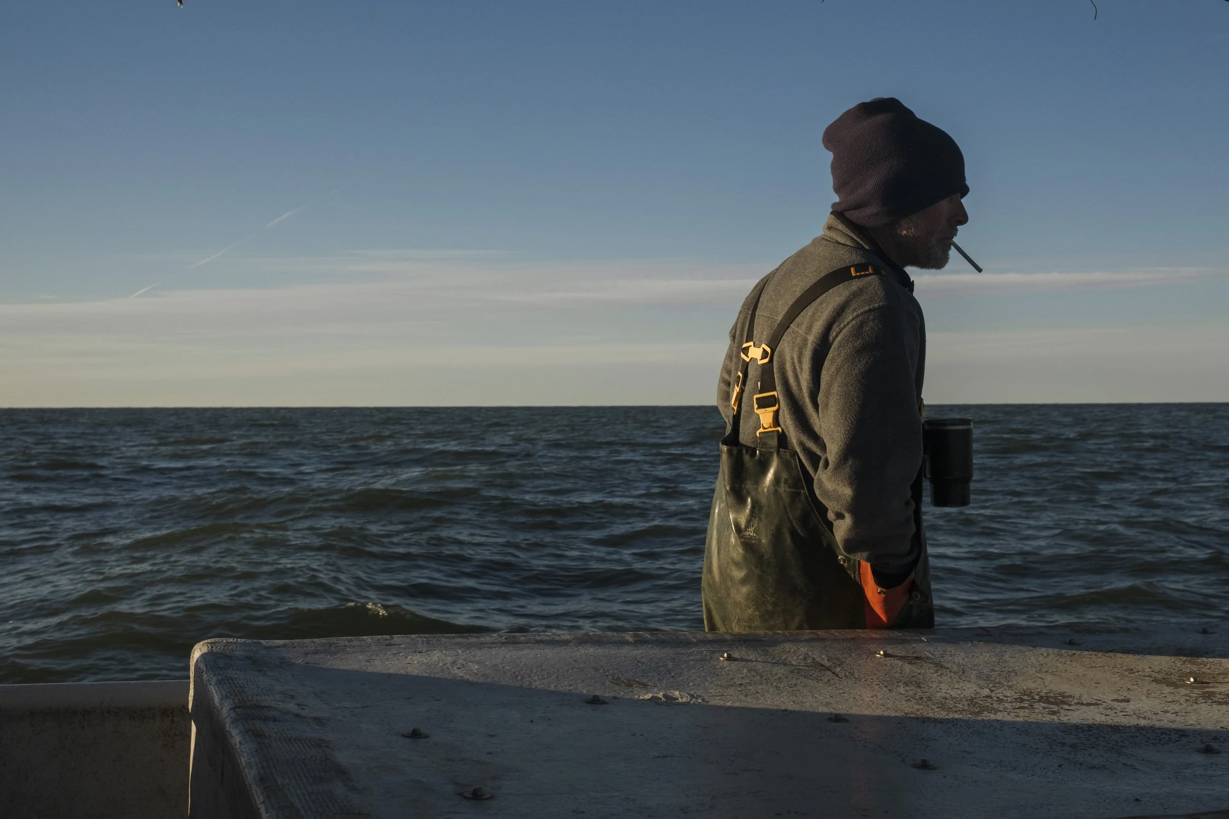 A man wearing a beanie, jacket, and gloves stands on a boat by the water, holding a cup with a view of the ocean and a blue sky.
