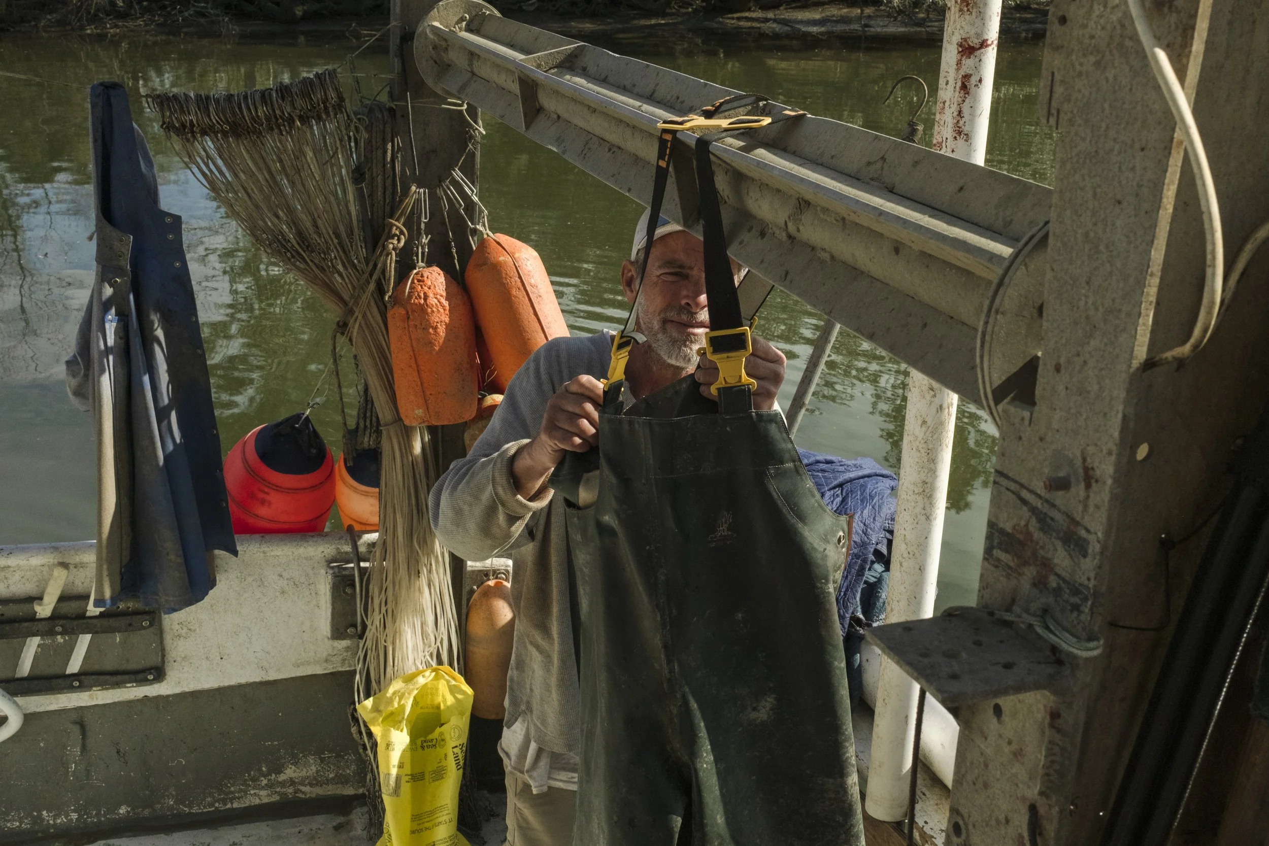 A man working on a boat, surrounded by fishing and boating equipment including buoys, a rake, and a ladder, with water in the background.