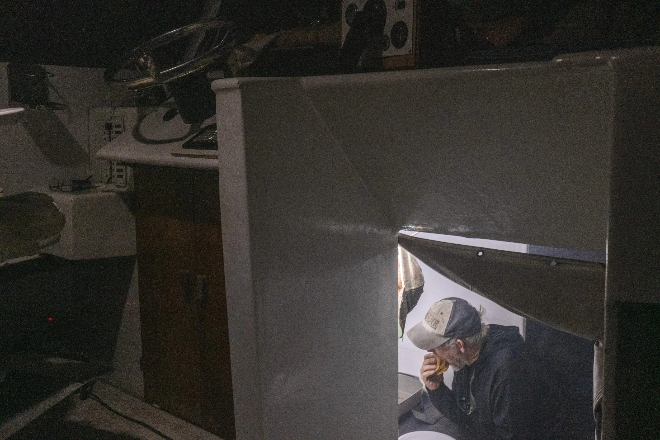 Man wearing a baseball cap eating a burger inside a small, enclosed space that resembles a kitchenette or food stall, seen through an opening in a partition.