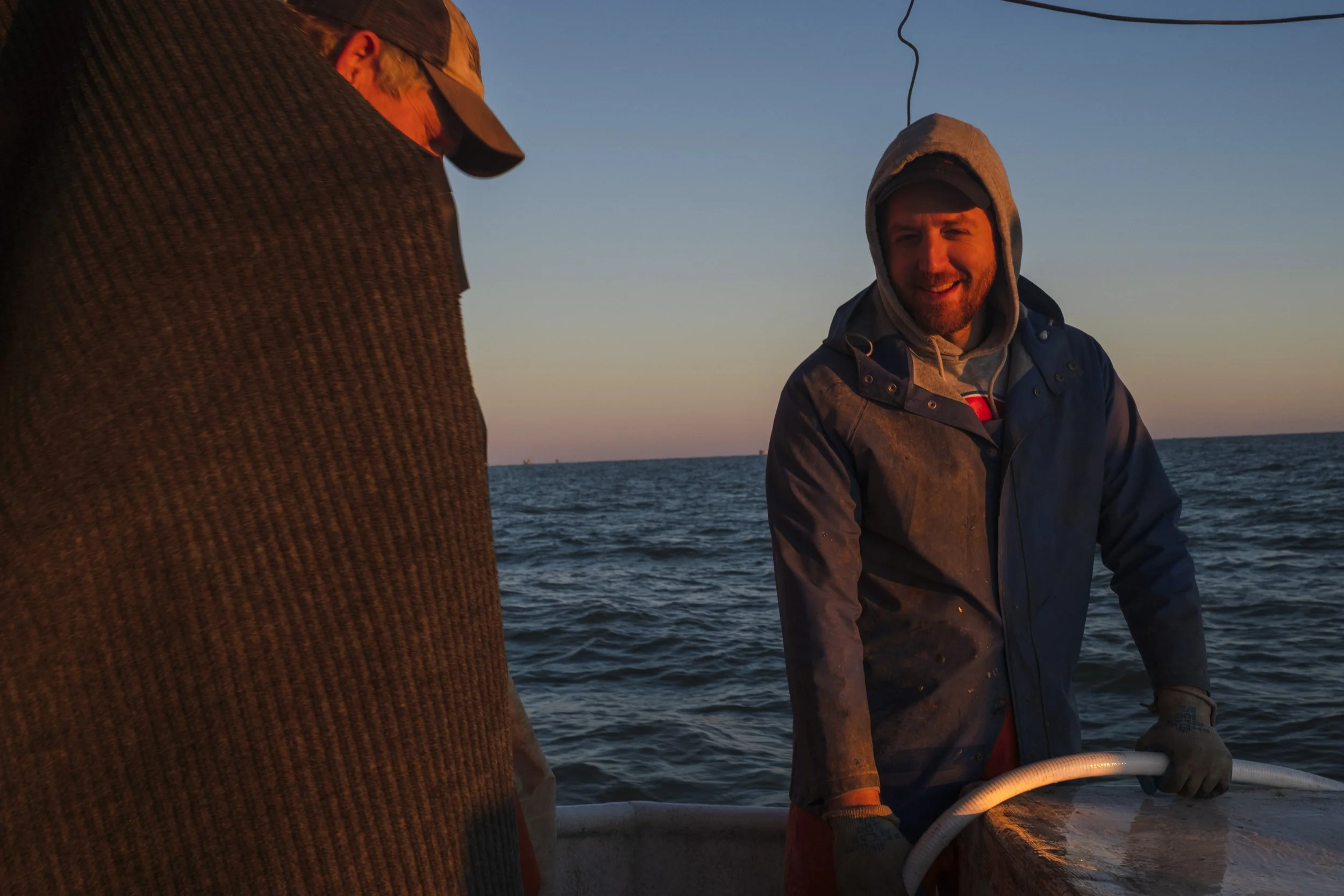 Two men on a boat at sunset, one smiling and wearing a blue jacket with a hood, and the other wearing a cap and a dark sweater, with the ocean in the background.