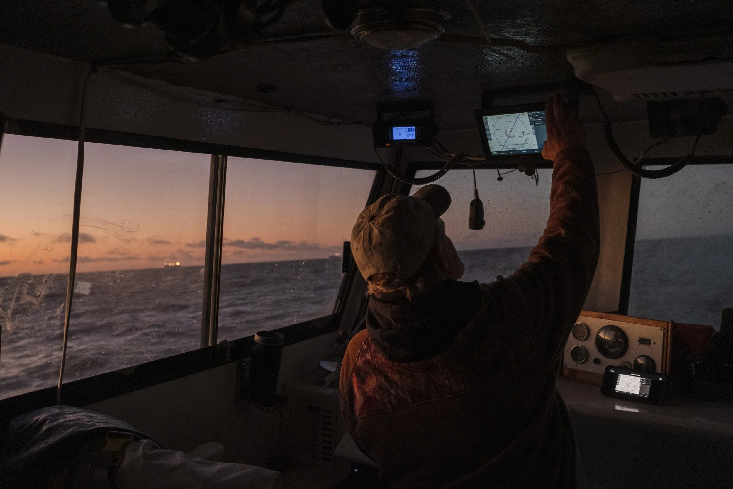 A person in a boat’s control room at sunset, wearing a baseball cap and using a navigation screen.