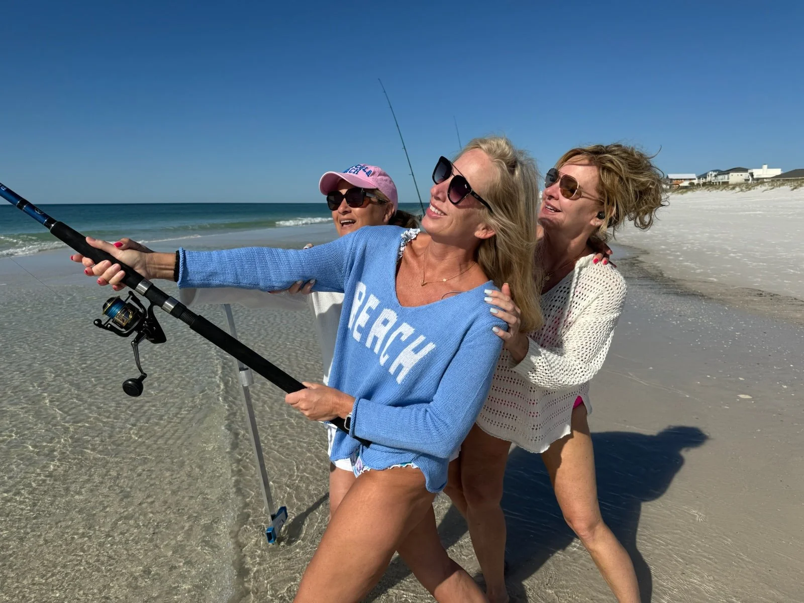 Three women standing on a sandy beach, posing for a photo with a fishing rod, with the ocean and clear blue sky in the background.