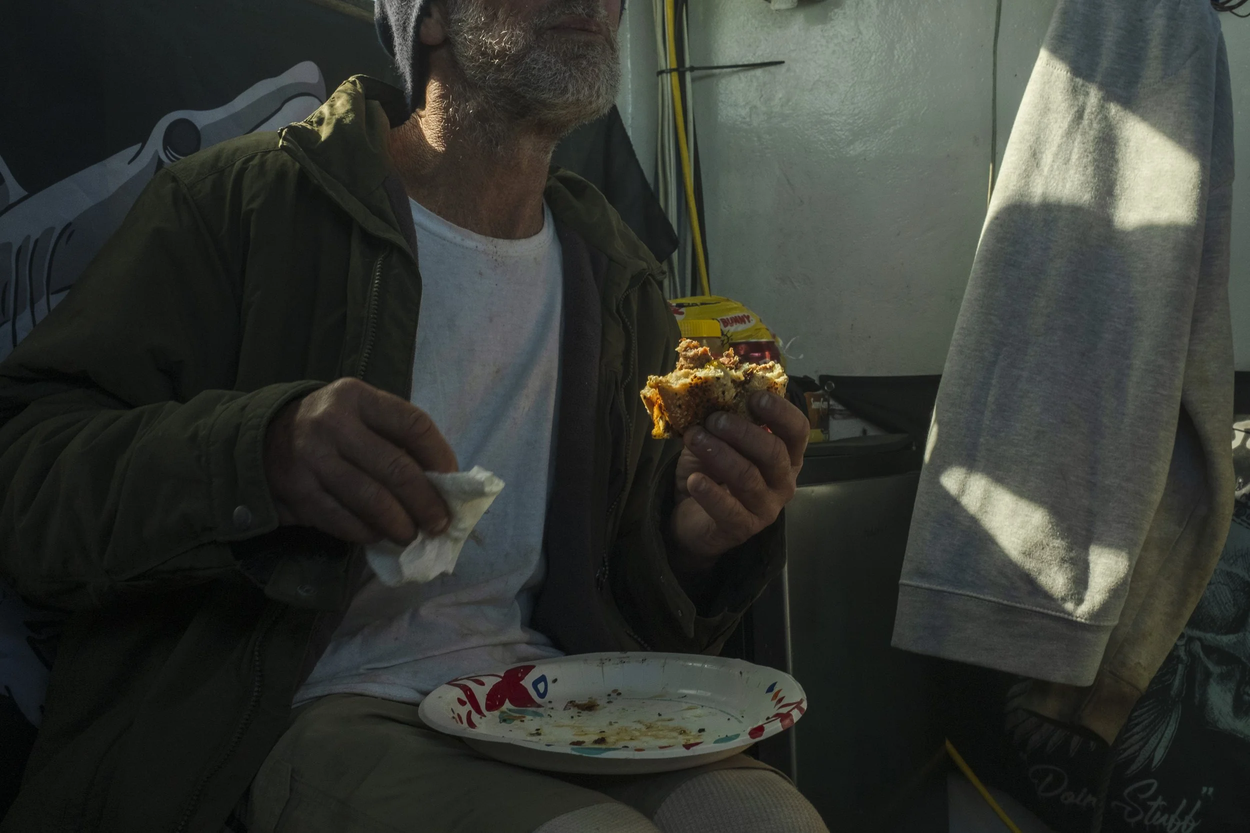 An older man with a gray beard wearing a dark jacket and white shirt, sitting down and holding a partially eaten piece of cake in one hand and a napkin in the other. In front of him, there's a disposable plate with crumbs on it. The setting appears t