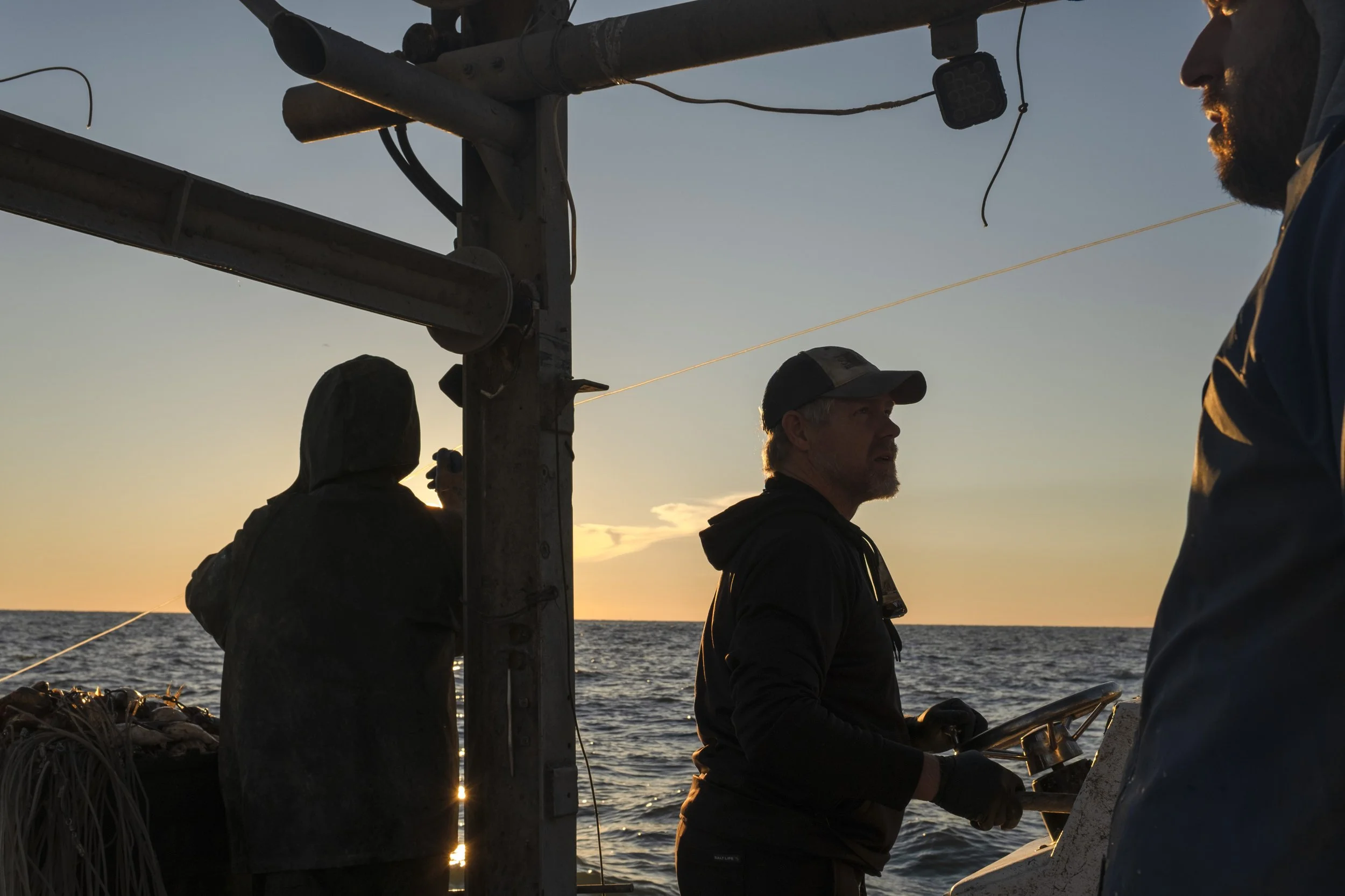 Three fishermen on a boat at sunset, one holding a fishing rod, with the ocean in the background.