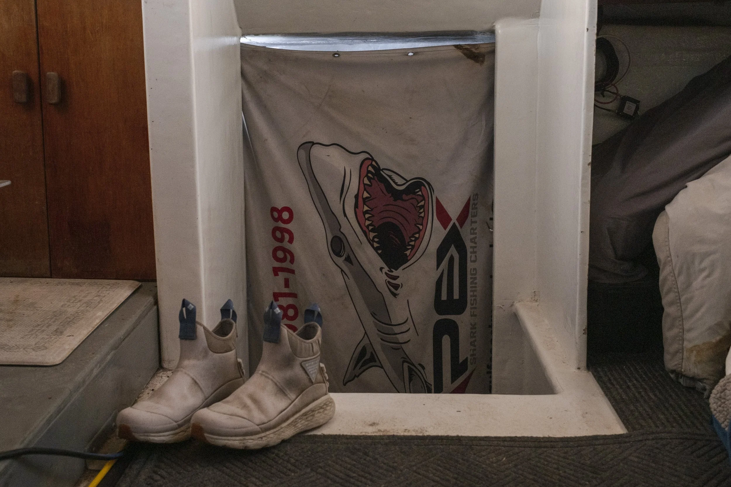 Pair of white shoes with blue tags on a black mat in front of a white opening. Inside the opening, there is a banner with a shark illustration and the text 'Starpark Fishing Charters' and a phone number.