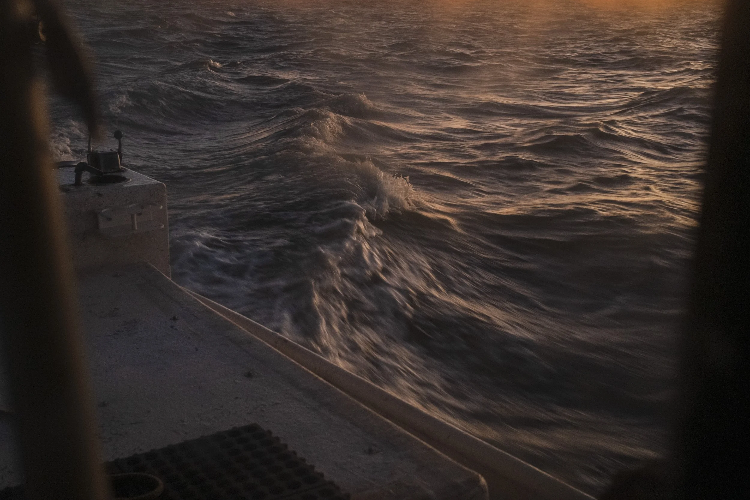 View of ocean water from a boat at sunset or sunrise, with ripples and waves, partially framed by the boat's structure.