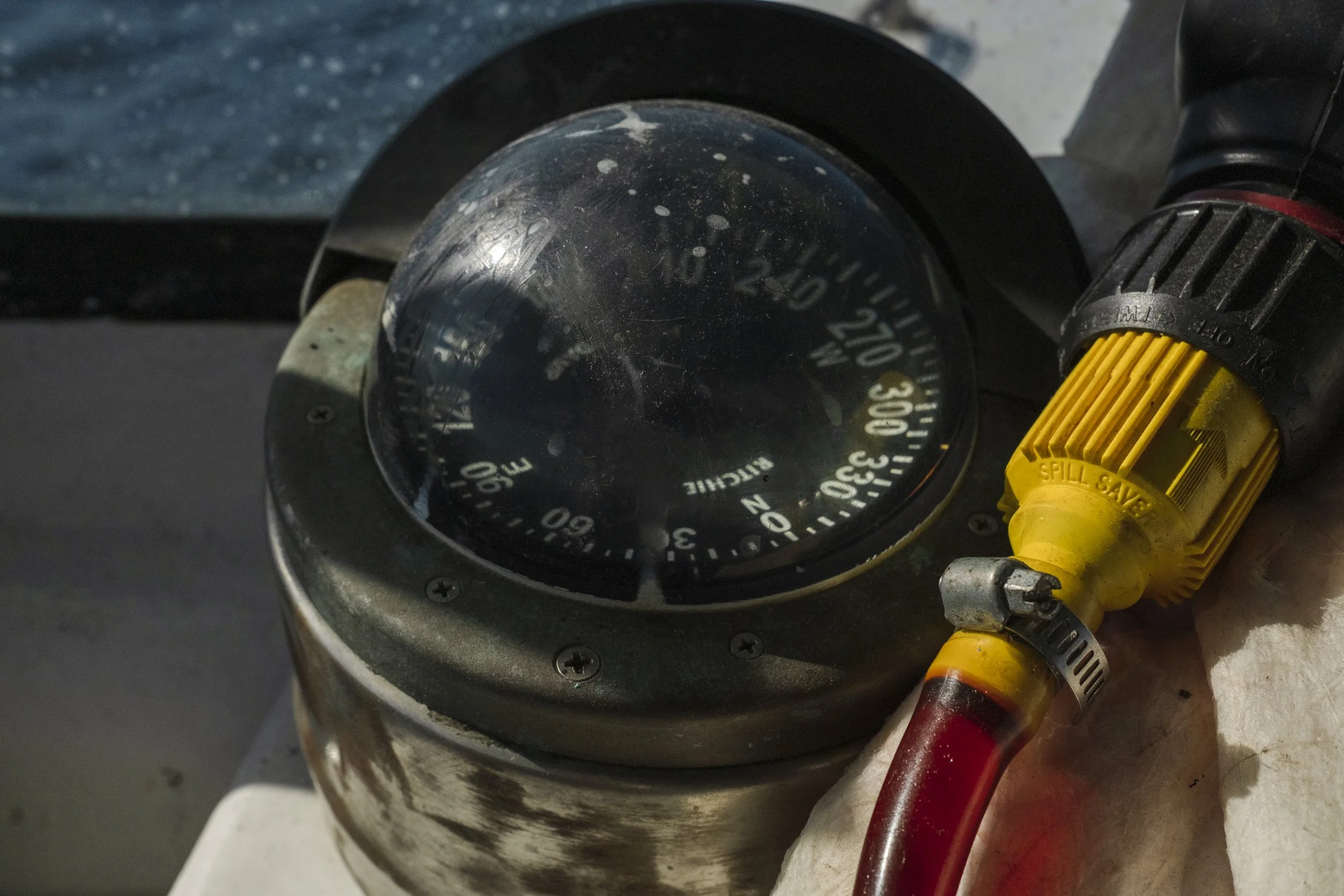 Close-up of a vintage dial stopwatch with a black glass face and white and yellow markings, accompanied by a yellow and black spill-saver valve connected to a red hose, resting on a white cloth surface.