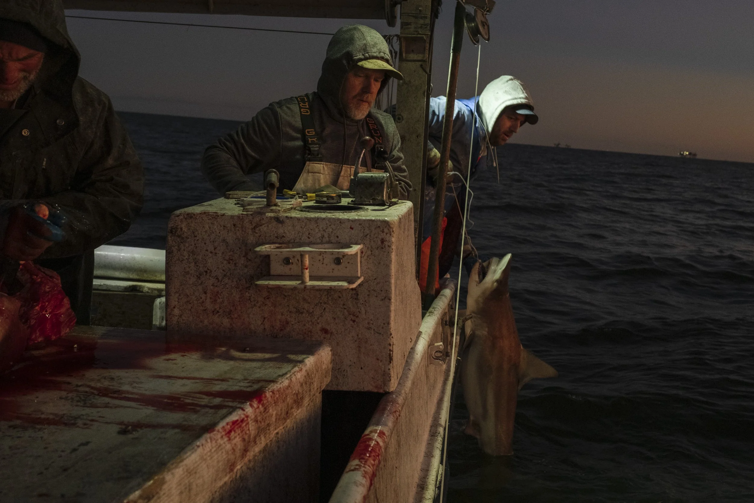 Two men on a boat at dusk, one holding a large fish, with another person partially visible. The boat has a weathered, blood-stained surface and the ocean stretches into the horizon with a ship visible in the distance.