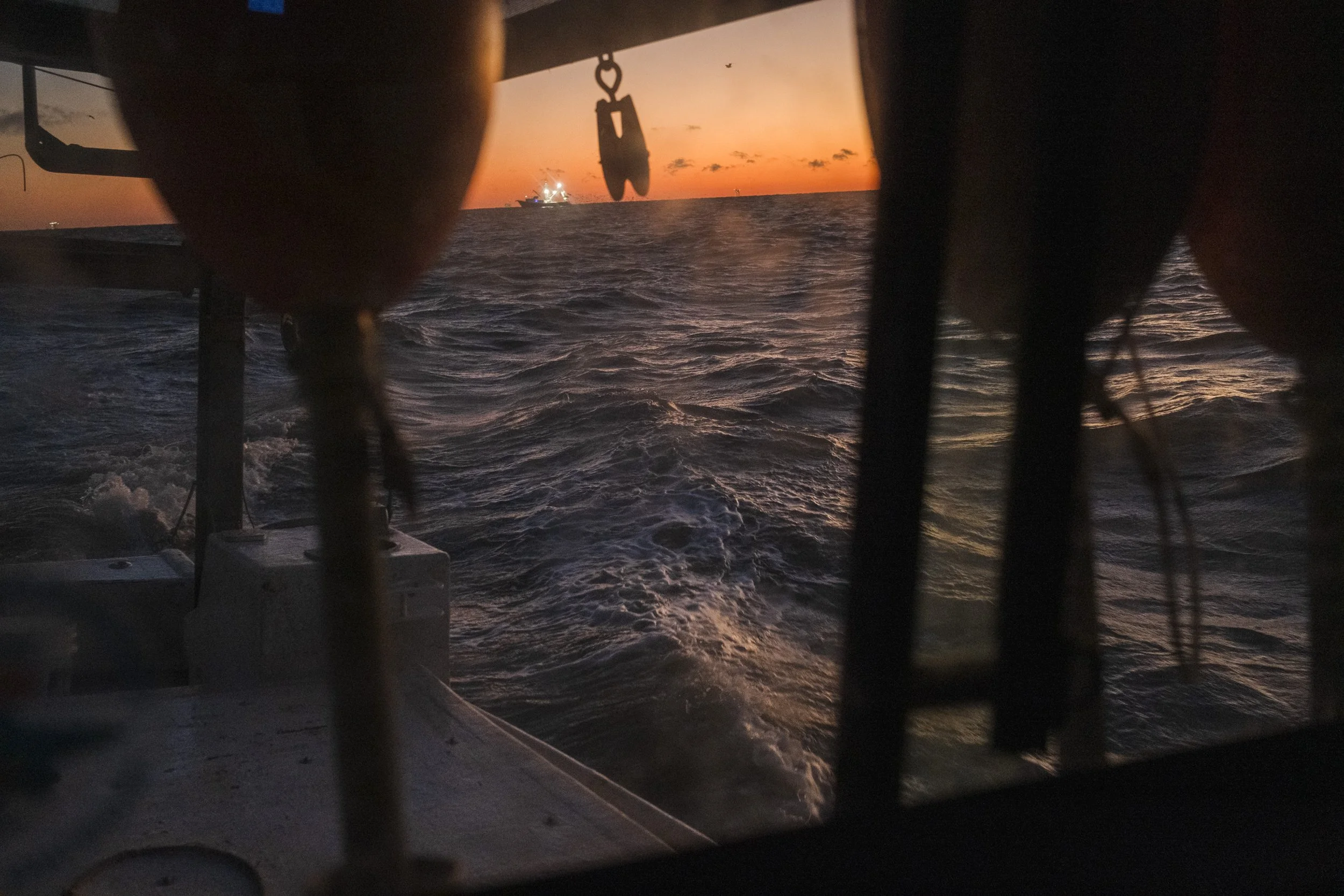 View from a boat at sunset with the ocean and distant ship in the background, framed by boat's structures and equipment.