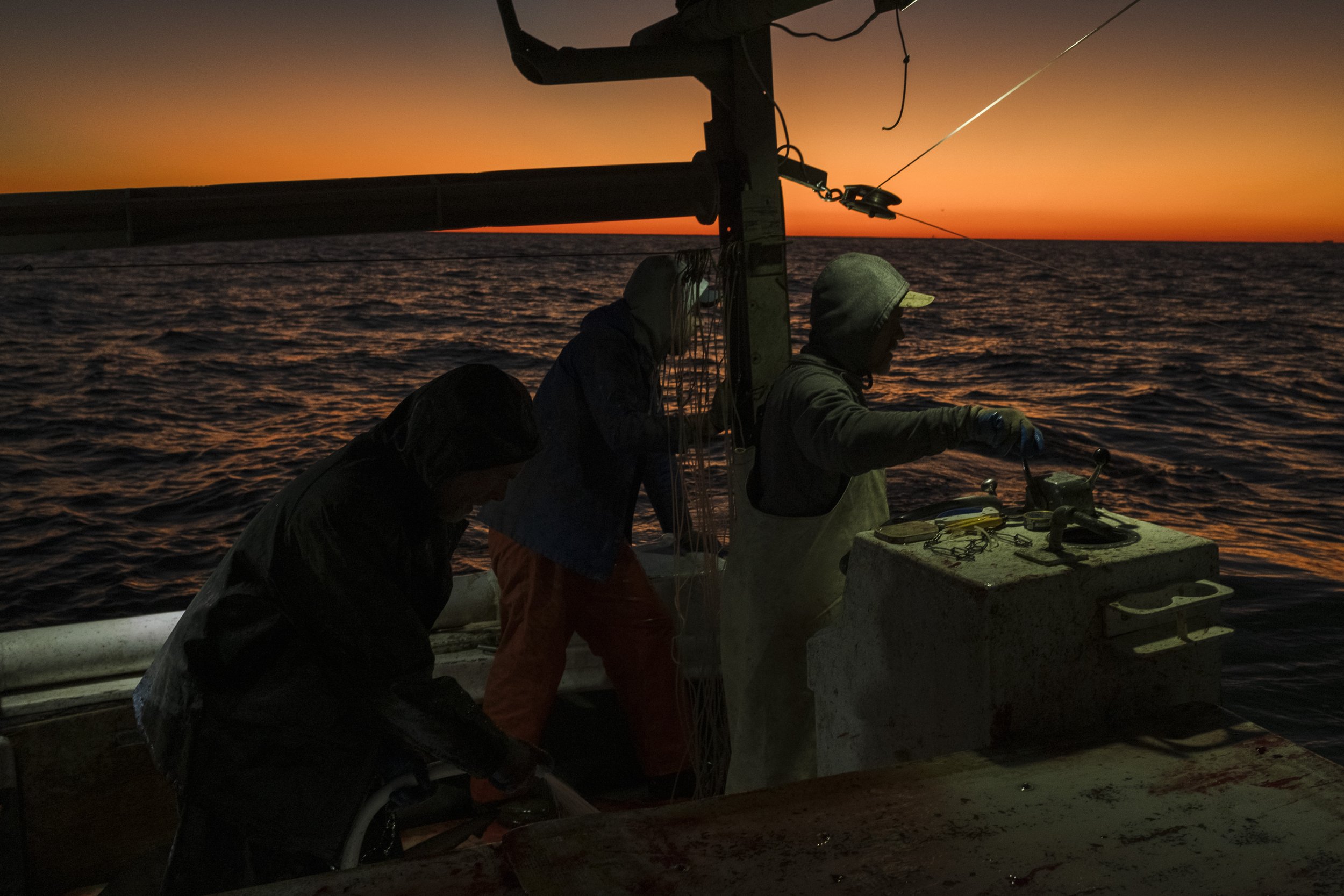 Three people working on a boat during sunset, with the ocean in the background.