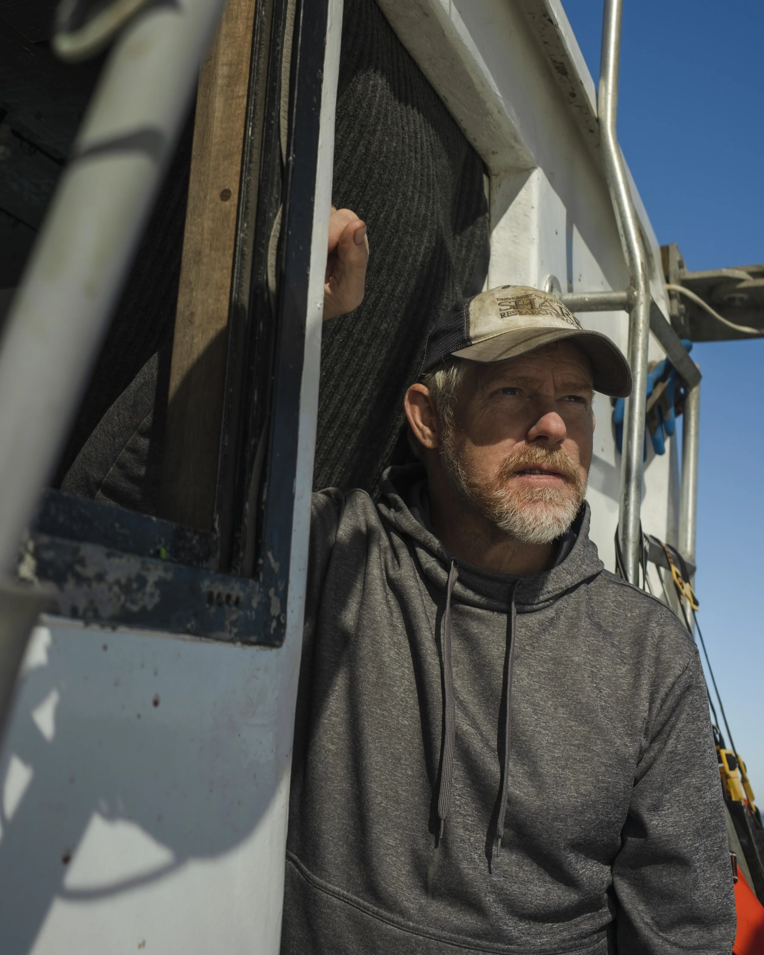 A man with a beard wearing a gray hoodie and a cap leaning out of a vehicle window, looking into the distance.