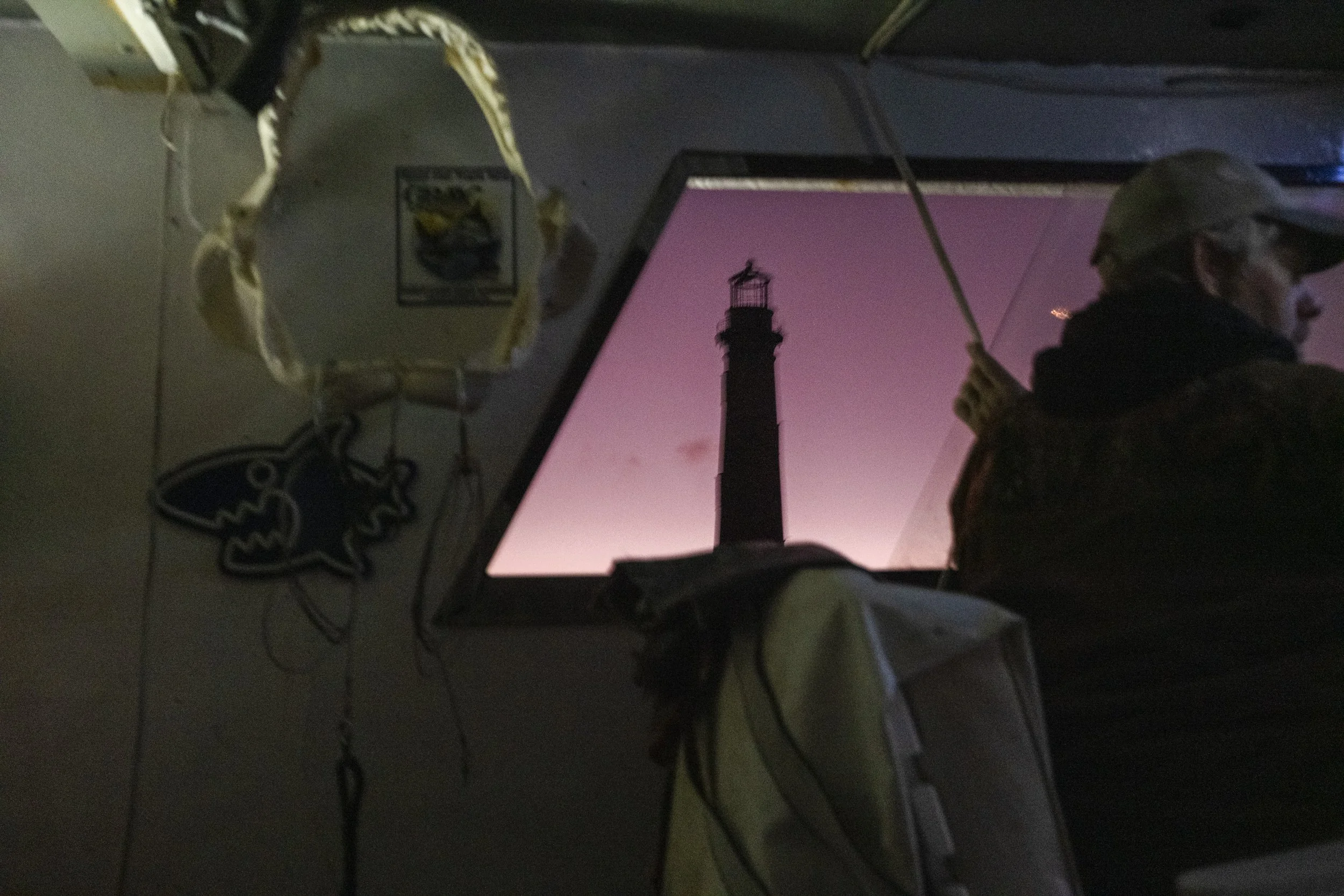 A person wearing a cap and glasses looking towards a computer monitor displaying a lighthouse silhouette against a purple sky, with various decorations hanging on the wall