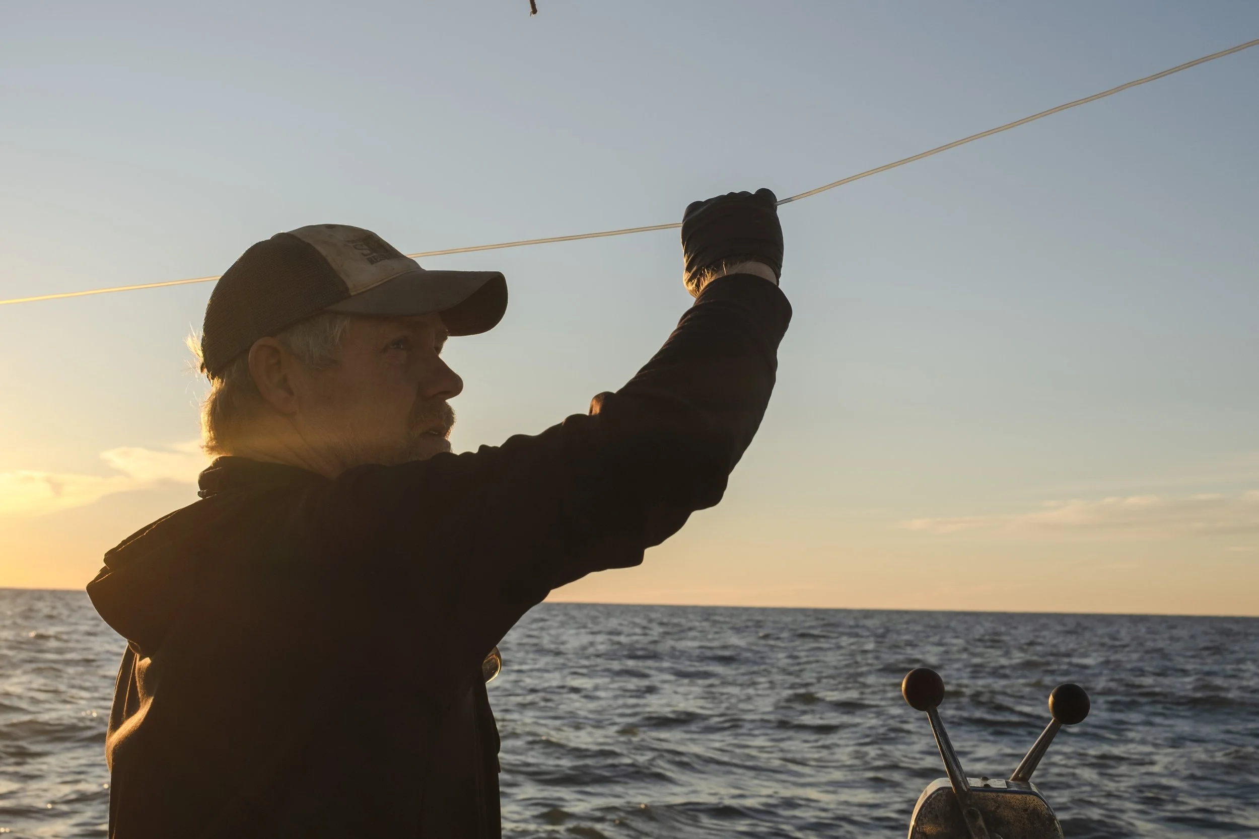 A man wearing a cap and dark clothing is fishing on a boat during sunset, holding a fishing line over the water.