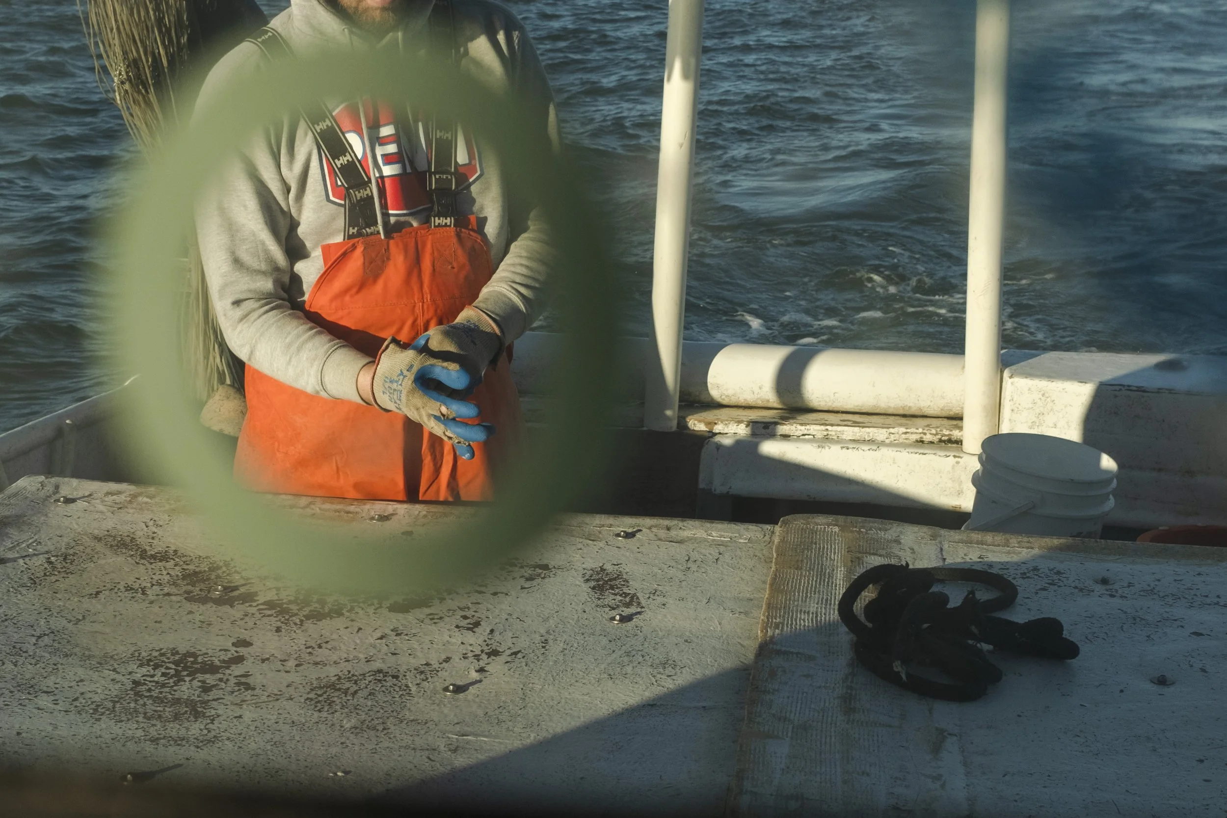 A person wearing orange waterproof bibs, a gray hoodie, and work gloves standing on a boat. The photo is taken through a green circular object, blurred in the foreground. The boat is on the water, with white railings and a bucket visible in the backg