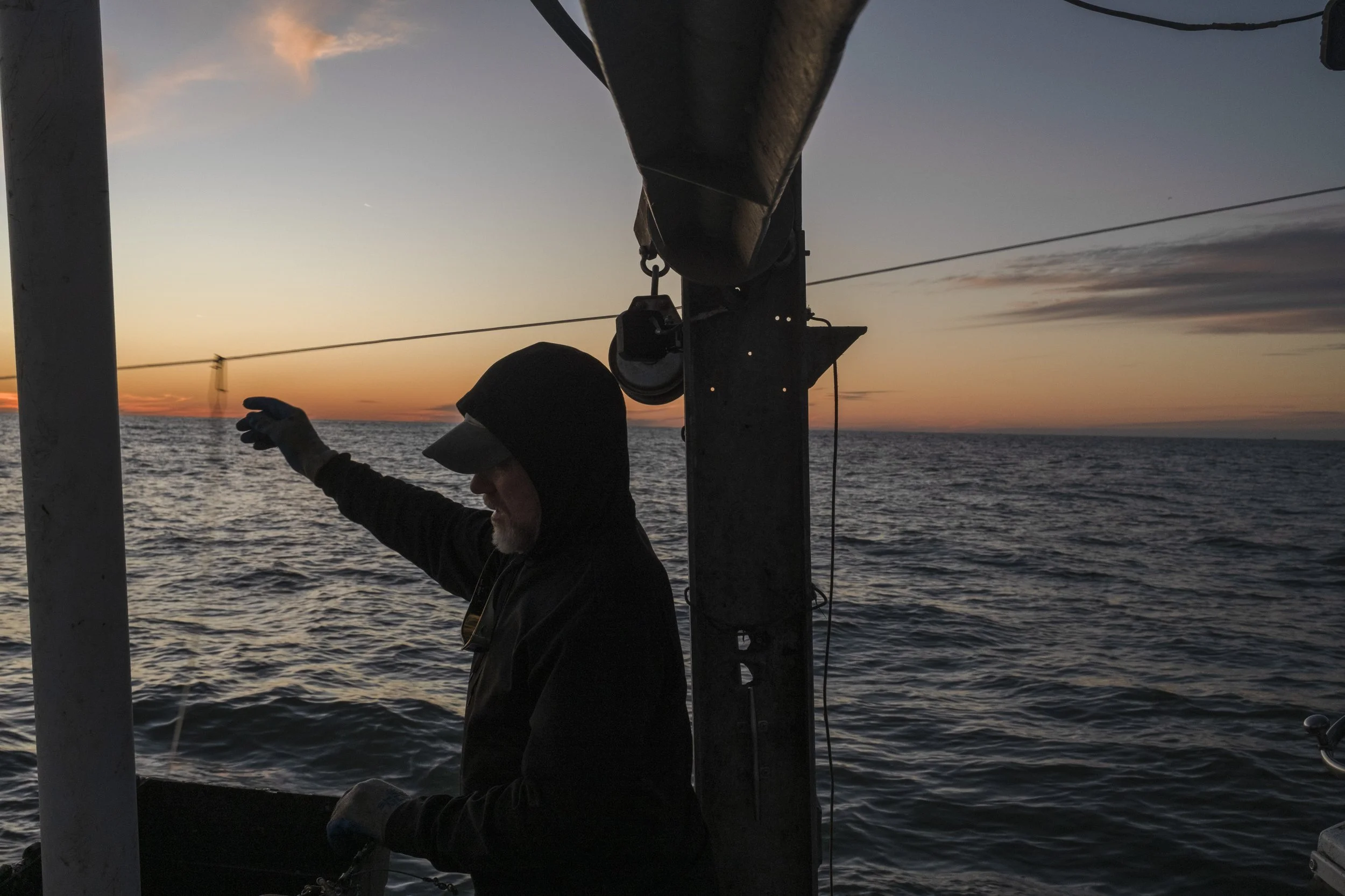 A person wearing a hoodie and gloves on a boat at sunset, reaching out over the water with a lines in the ocean and the horizon visible in the background.
