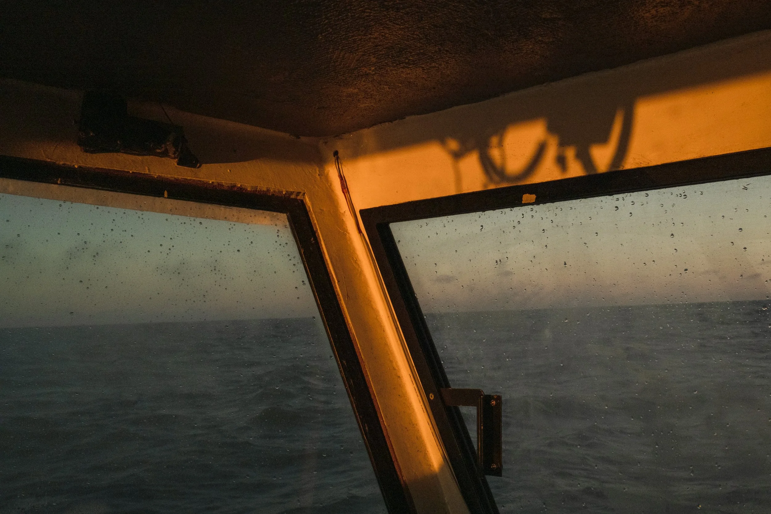 View from a boat's cabin showing rain-streaked windows with a sunset sky and calm ocean outside