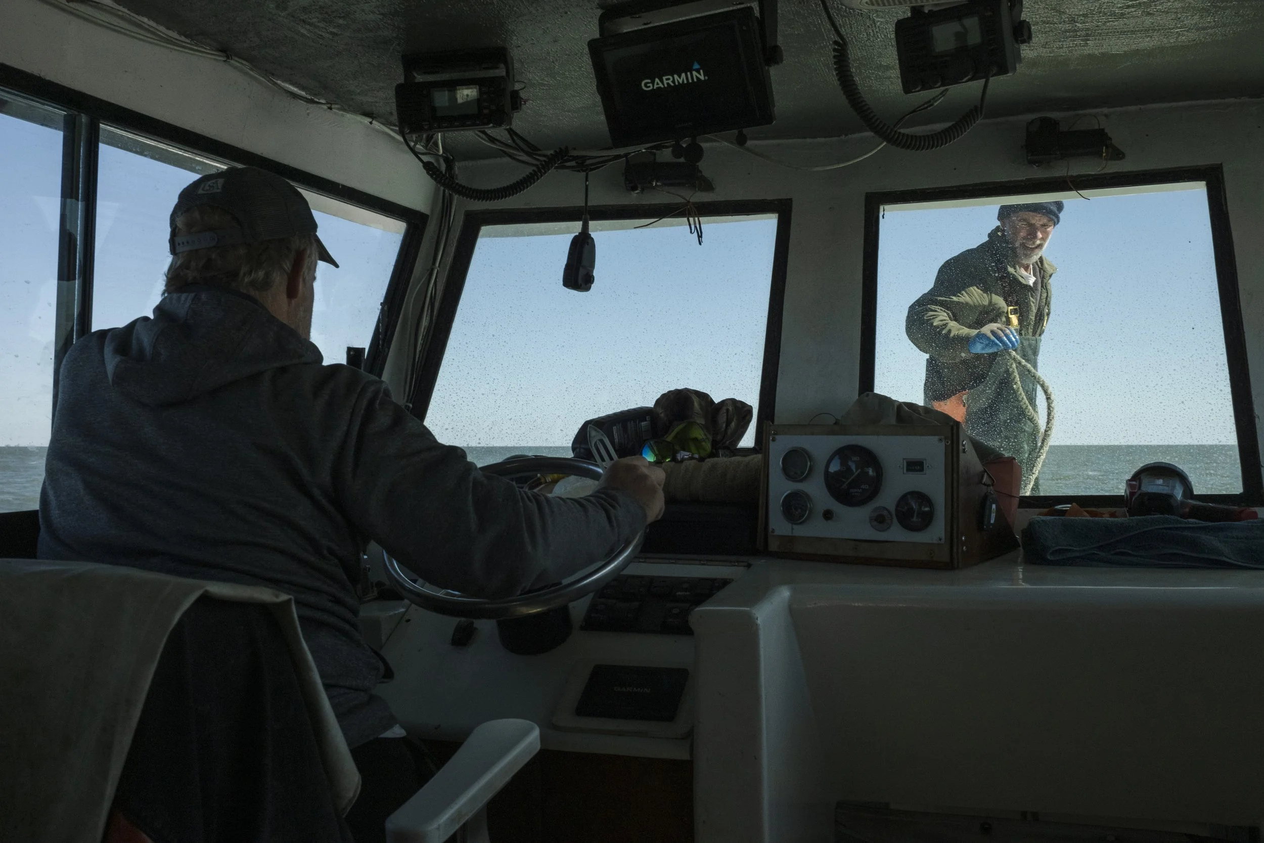 A person steering a boat with another person outside on the deck, handling a rope, seen through the boat's front window on a clear day.