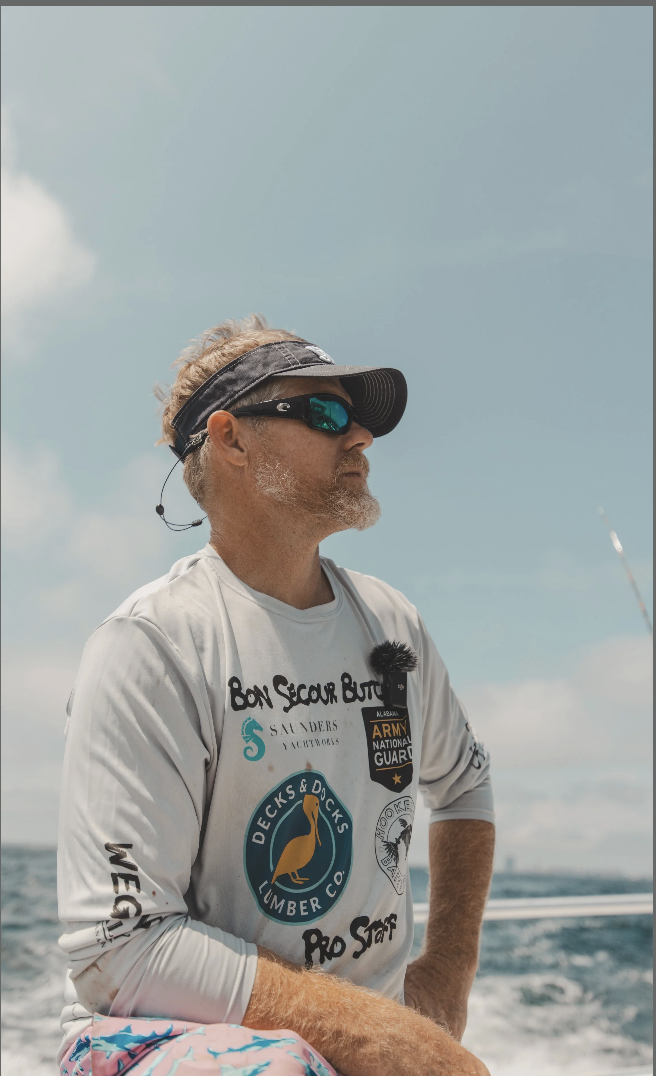 Man wearing sunglasses, a visor, and a long-sleeve shirt with logos on a boat in the water.