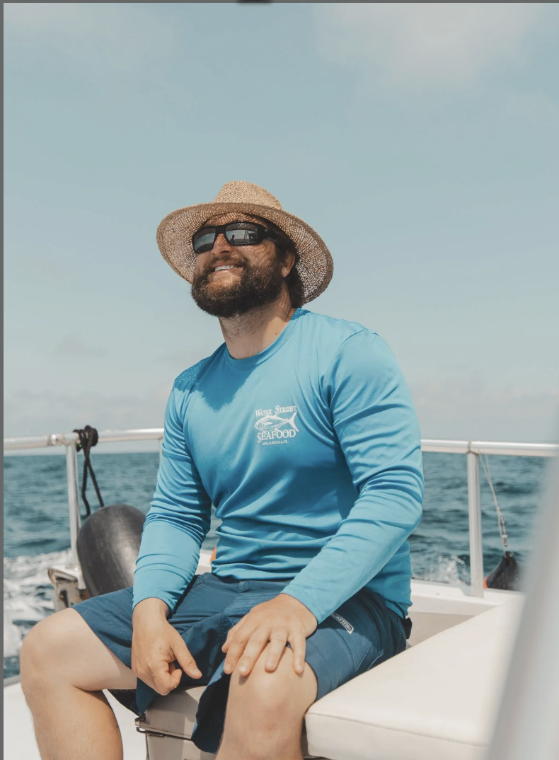A man with a beard and sunglasses sitting on a boat wearing a blue long sleeve shirt, a straw hat, and shorts, smiling as he enjoys a day out on the water.