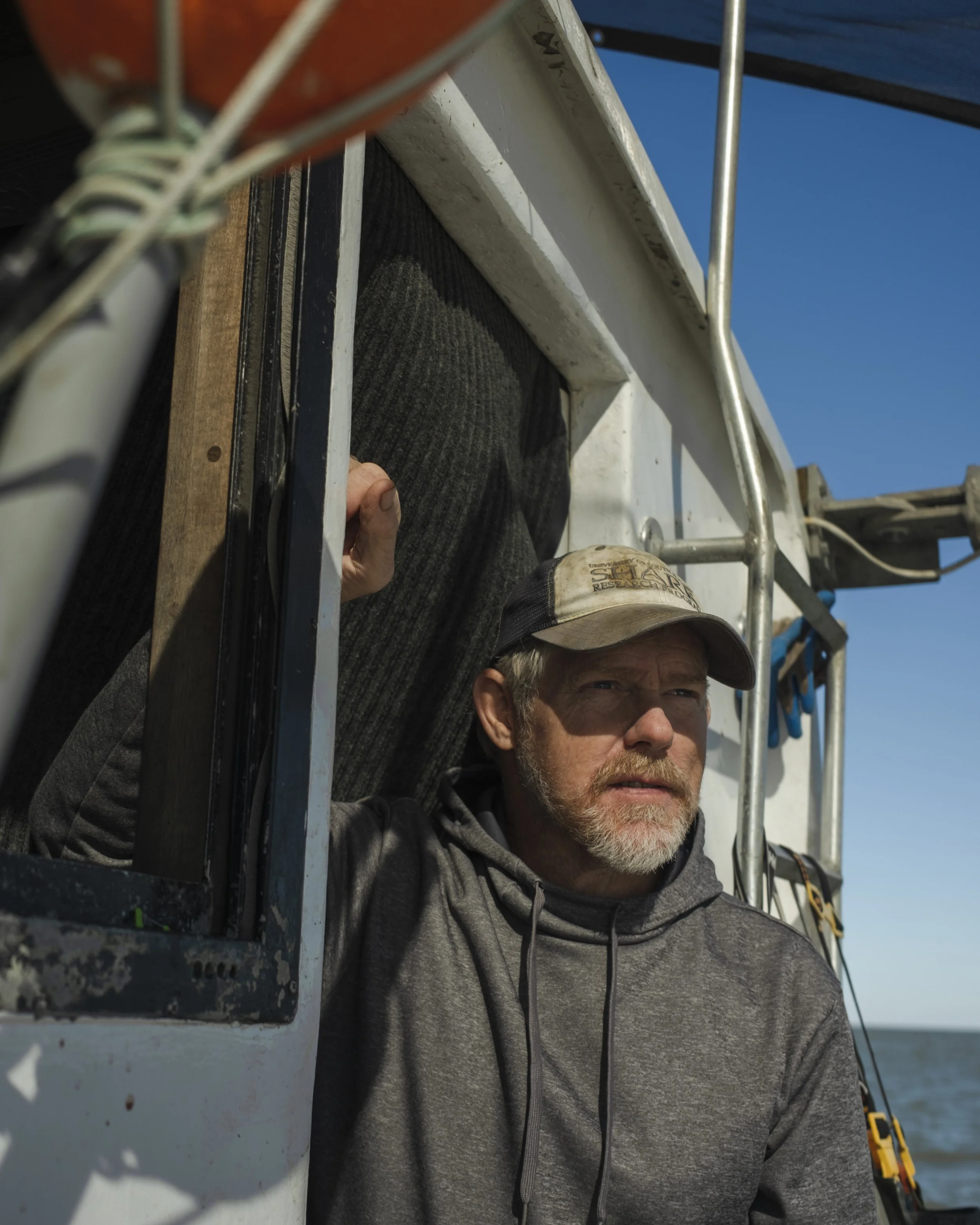 Man leaning out of a boat window, wearing a hoodie and cap, with water in the background.