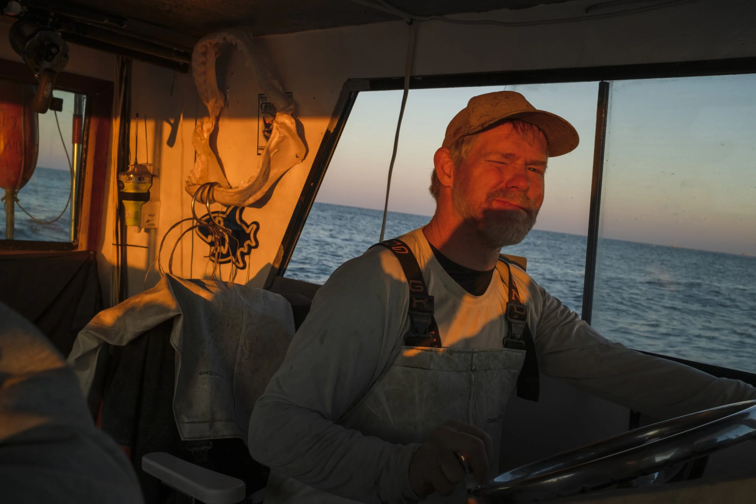 A man with a cap and beard steering a boat on the open water during sunset, with the sunlight illuminating his face and the boat's interior.