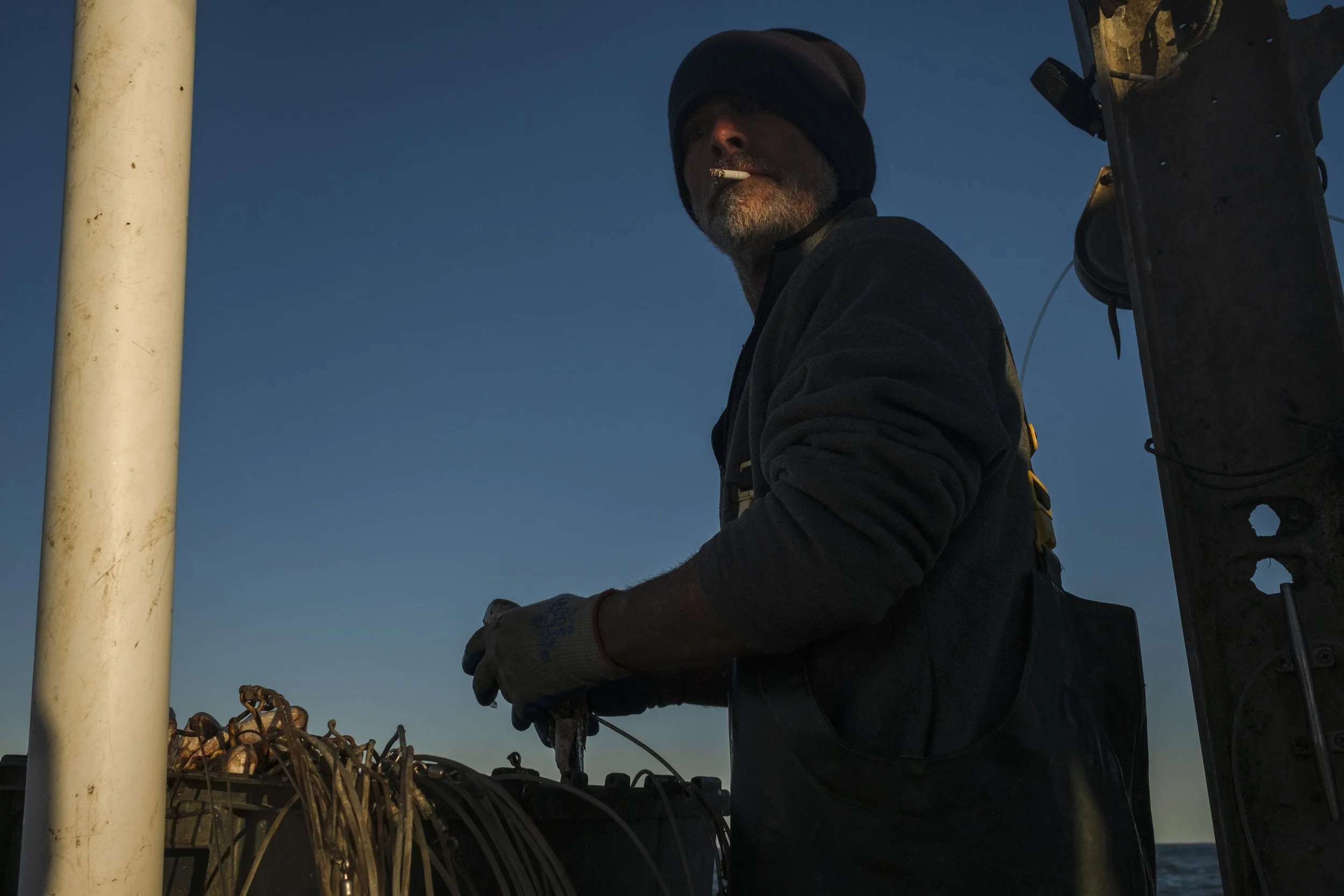 A man with a beard, wearing a black hoodie, gloves, and a beanie, stands on a boat during sunset, with the ocean and a clear sky in the background, and appears to be working with fishing equipment.