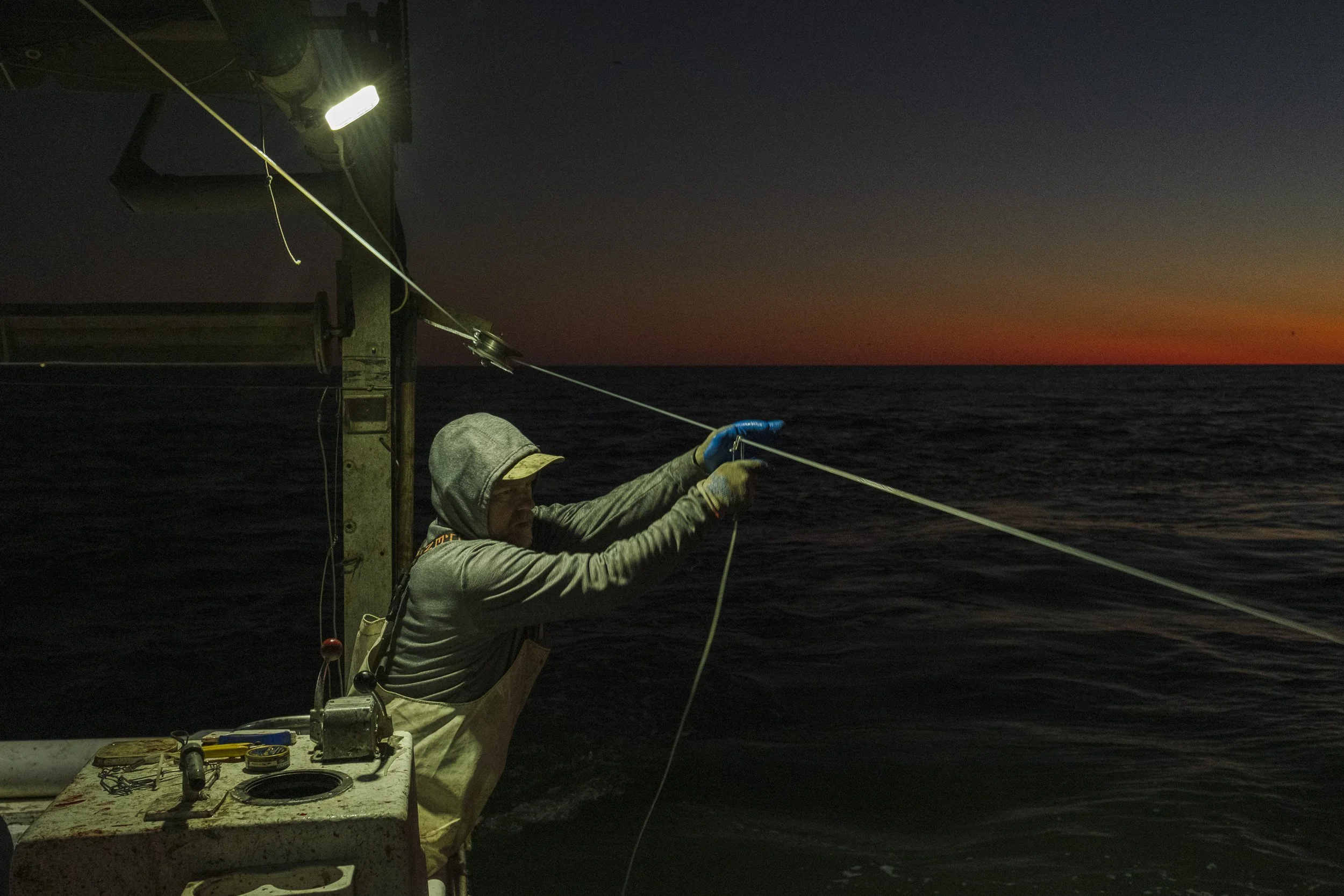 A person fishing off a boat at sunset, equipped with gloves and a hoodie, standing by a table with fishing tools.