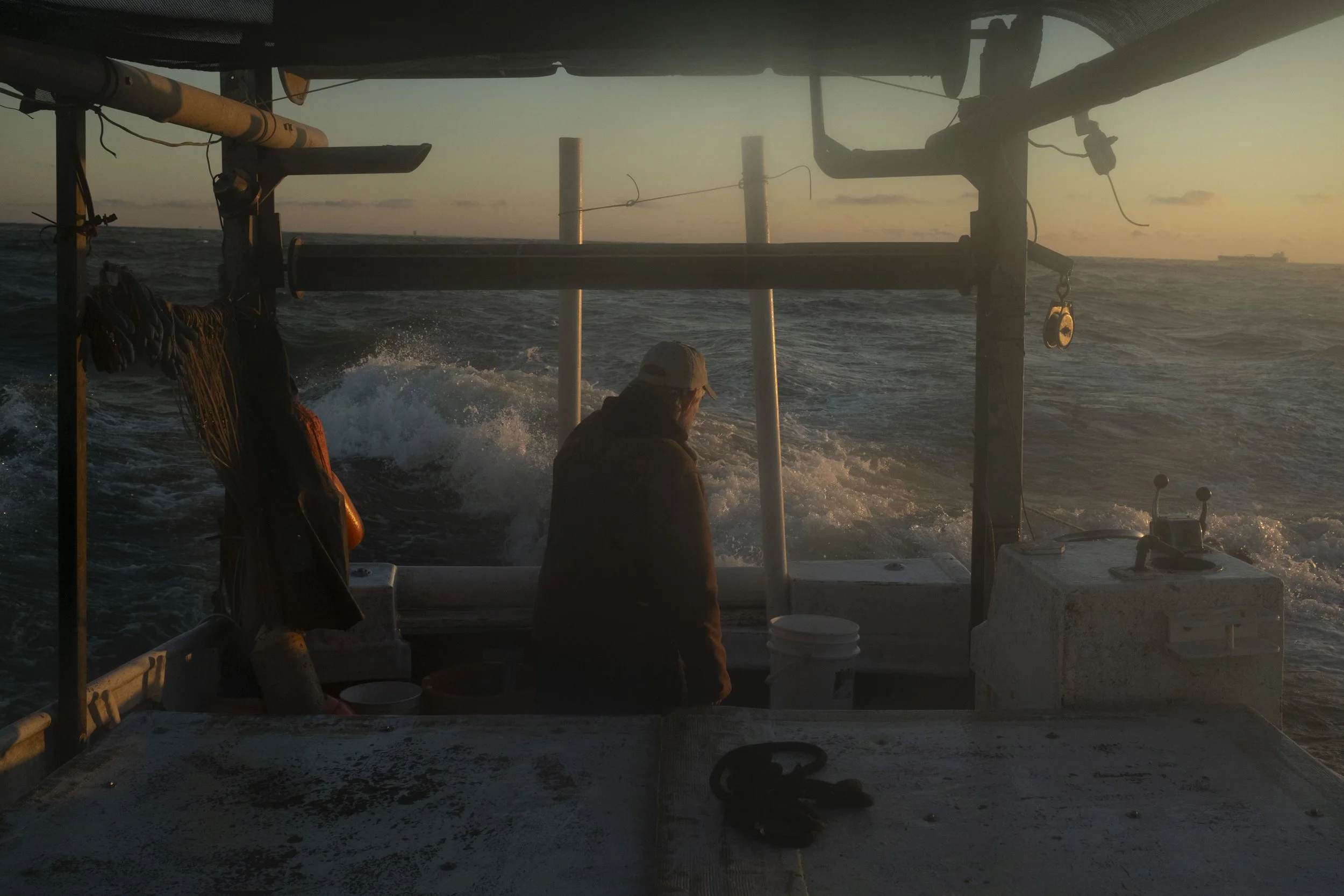 A person standing on a boat during sunset, with ocean waves crashing behind them, wearing a cap and dark jacket.