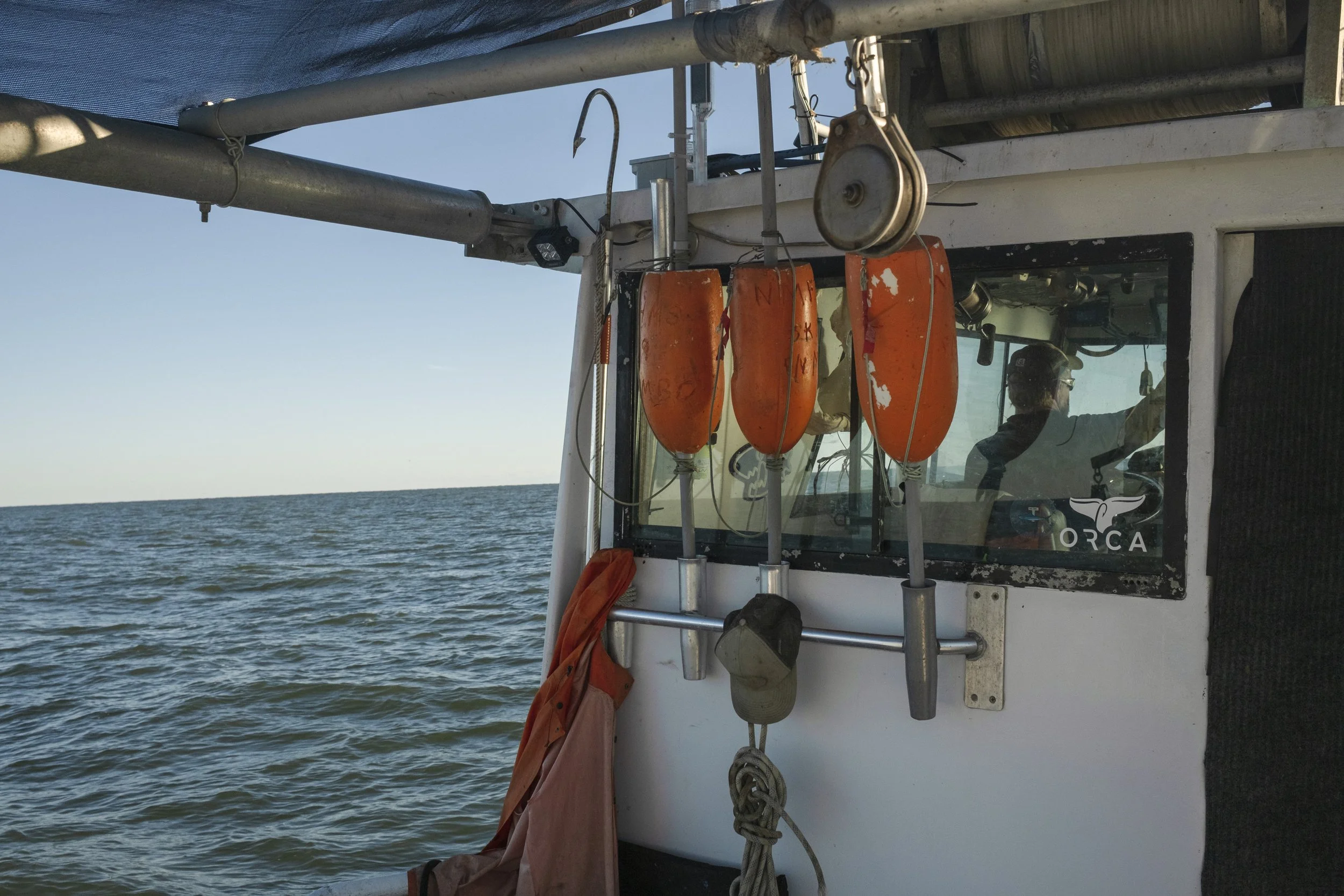Close-up of a boat's cabin with three orange floatation devices hanging on the side, a fishing hook and pulley, a cap, and a person with sunglasses inside the cabin steering the boat. The ocean and blue sky are visible in the background.