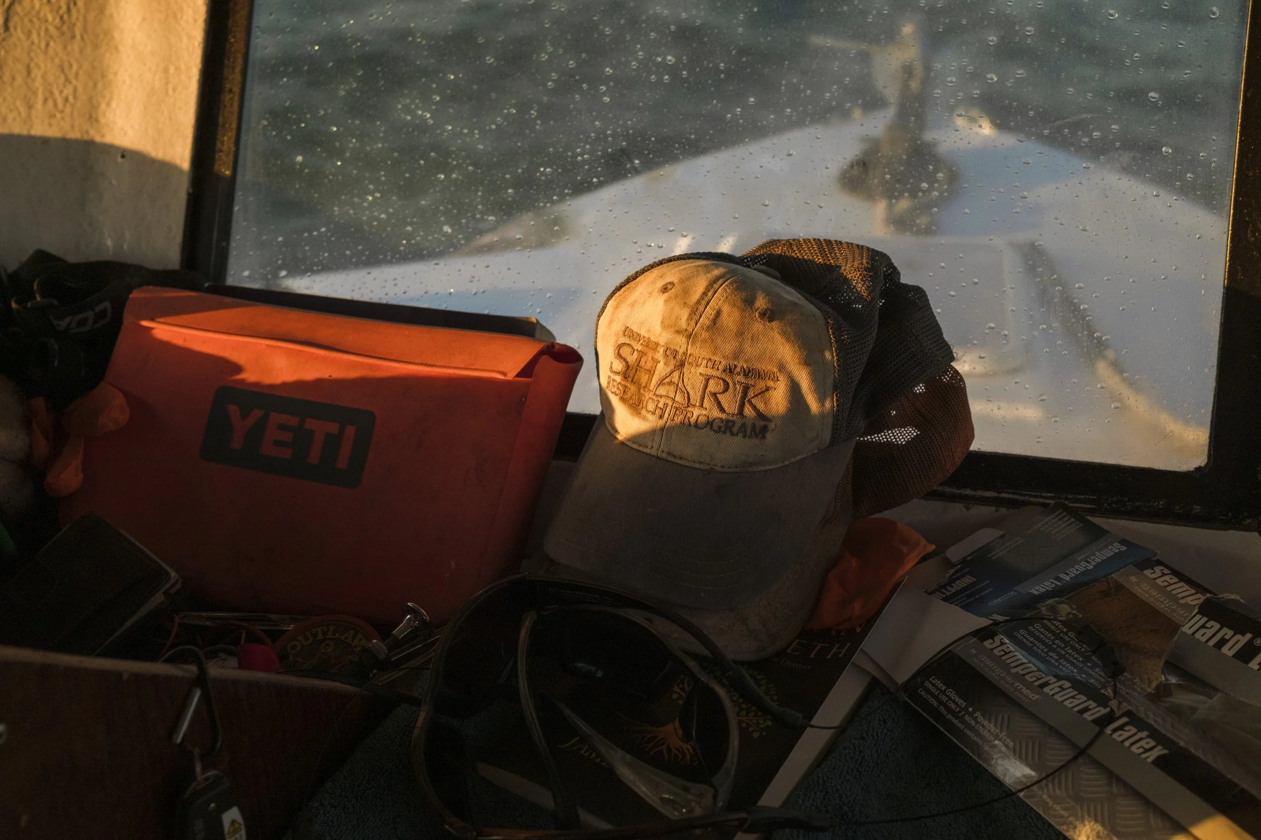 Inside a boat cabin, a YETI cooler, a baseball cap with 'SHARK' embroidered, sunglasses, and various magazines and supplies on a surface in front of a window showing water with raindrops.