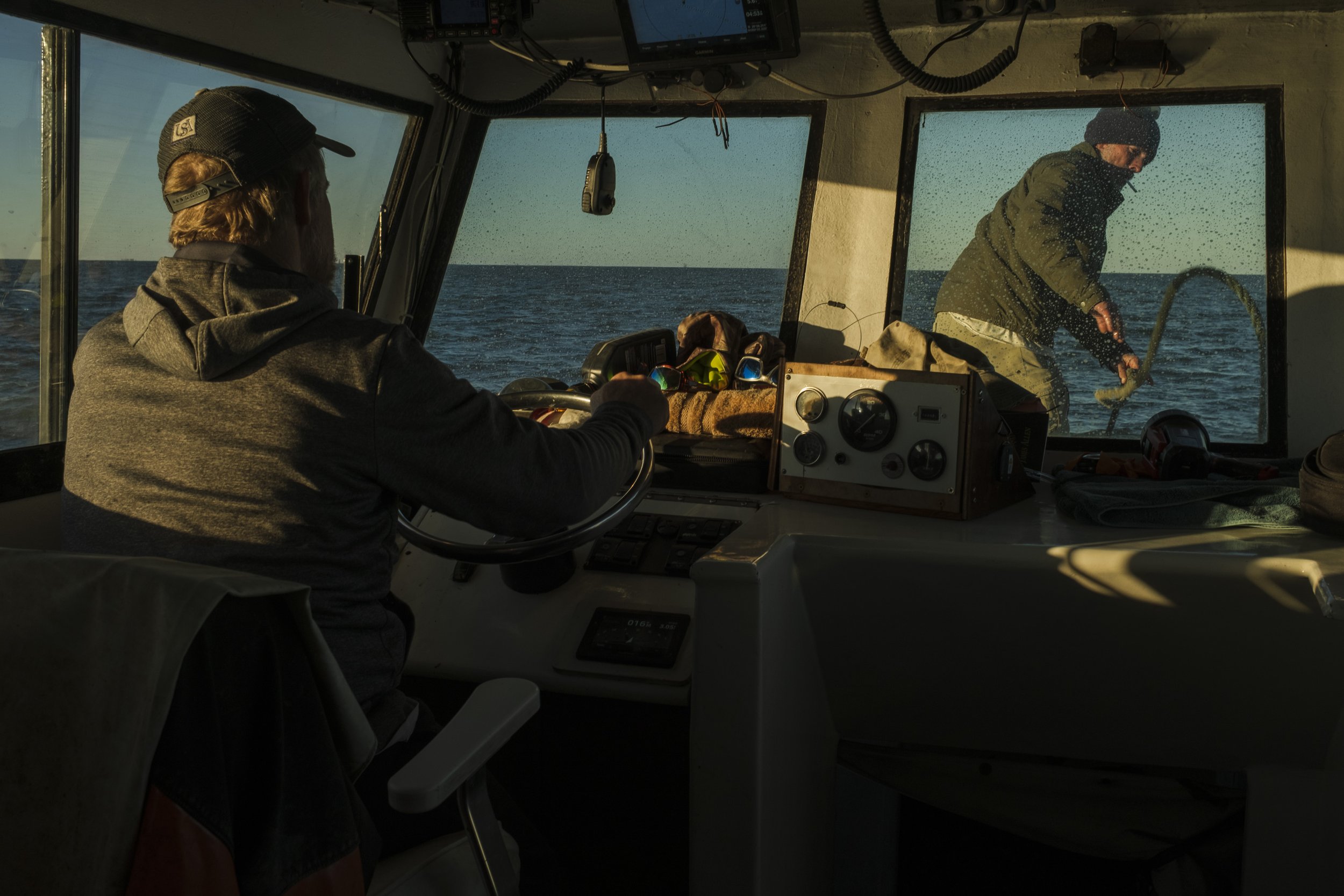 A person steering a boat on the water, with another person on the deck or dock outside the window, holding an object in the water. The boat's interior contains various controls and a collection of sunglasses, with ocean scenery visible through the wi