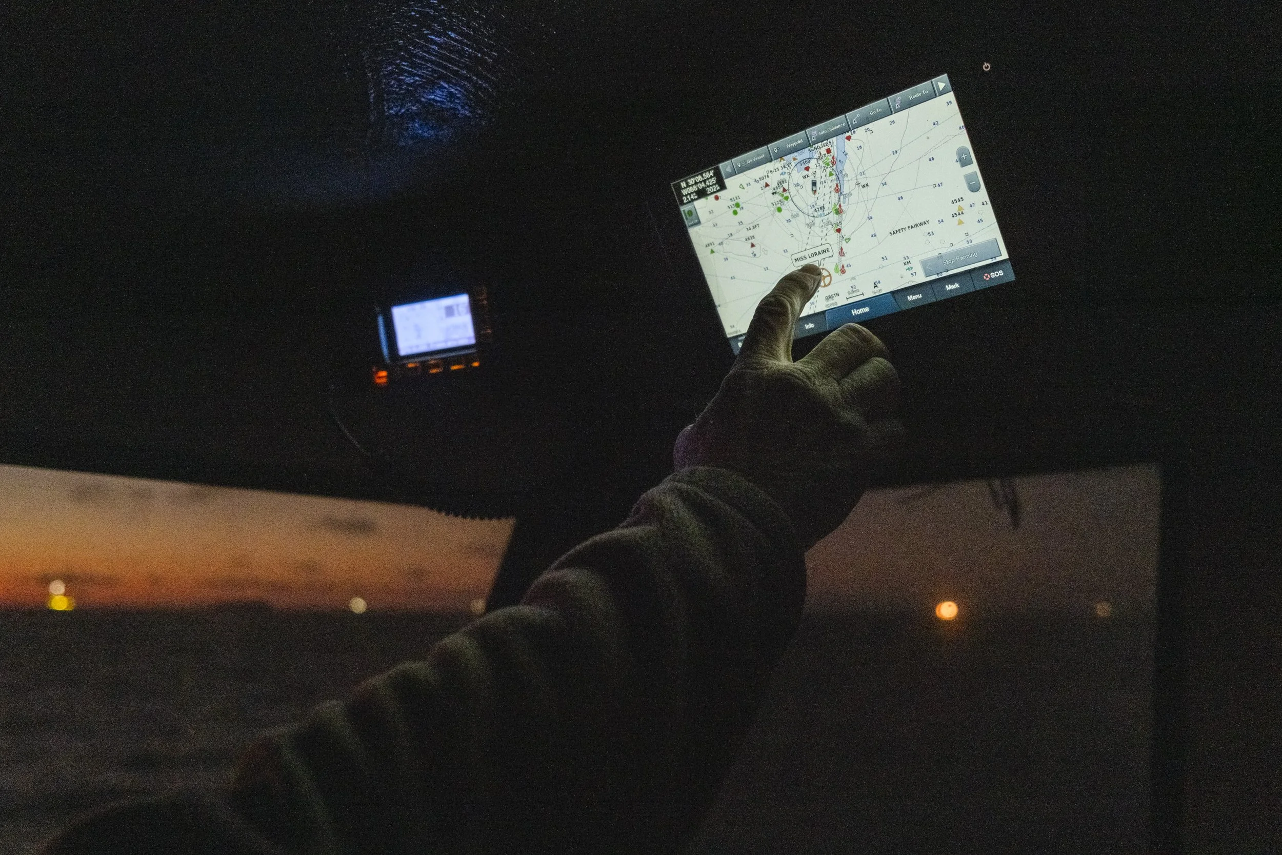 Person using a touchscreen navigation system inside a vehicle at dusk or night, with a sunset visible through the window and a small electronic device nearby.