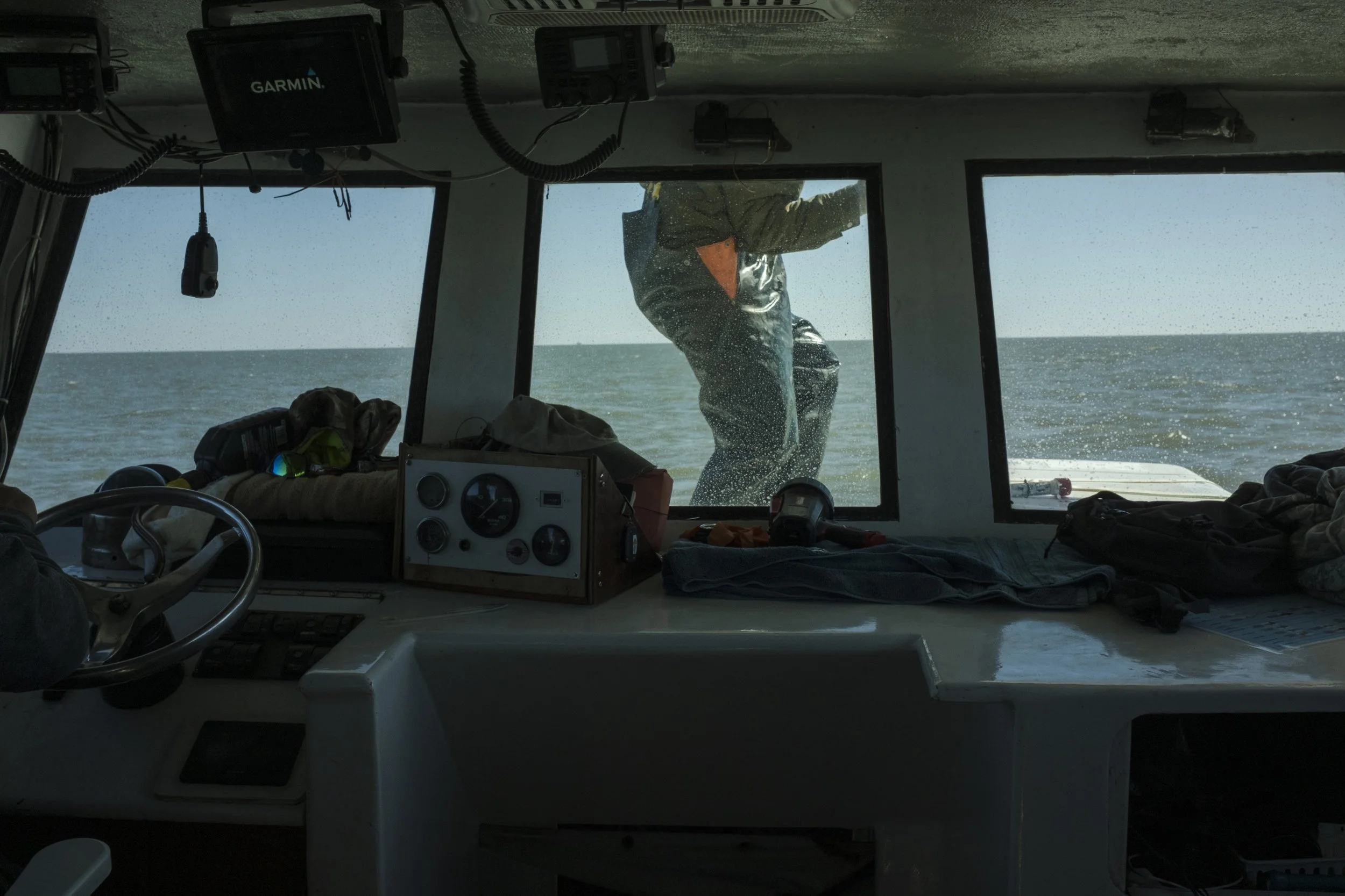 View from inside a boat cabin showing a person standing outside on the boat deck. The cabin has large windows, various navigation and communication devices, and some clothing and equipment on the interior surface.