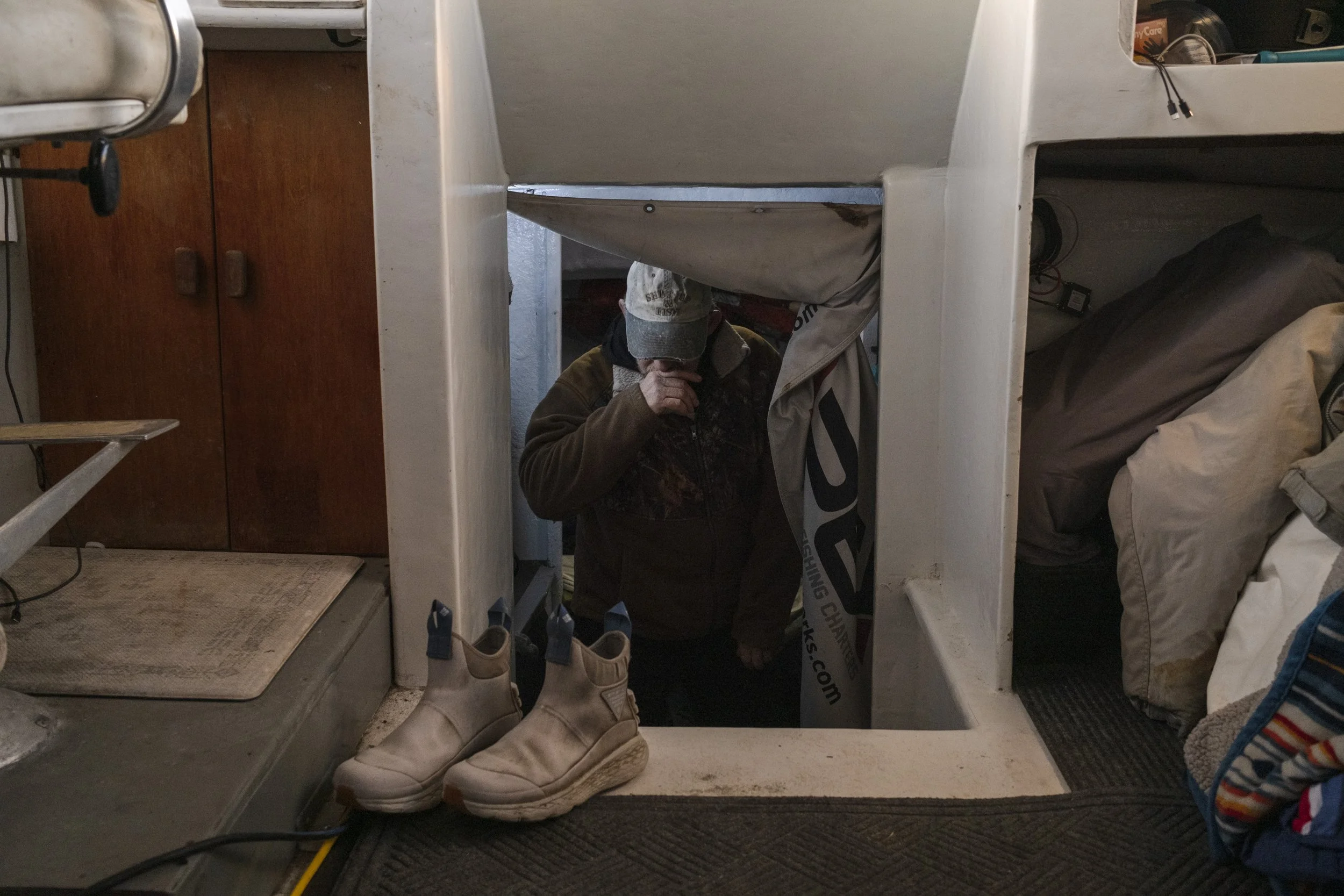Person wearing a cap and brown jacket crouching in a basement or storage area, partially visible through a small opening with a curtain. Shoes are placed on a lip at the entrance, with a cluttered room with sleeping bags and boxes in the background.