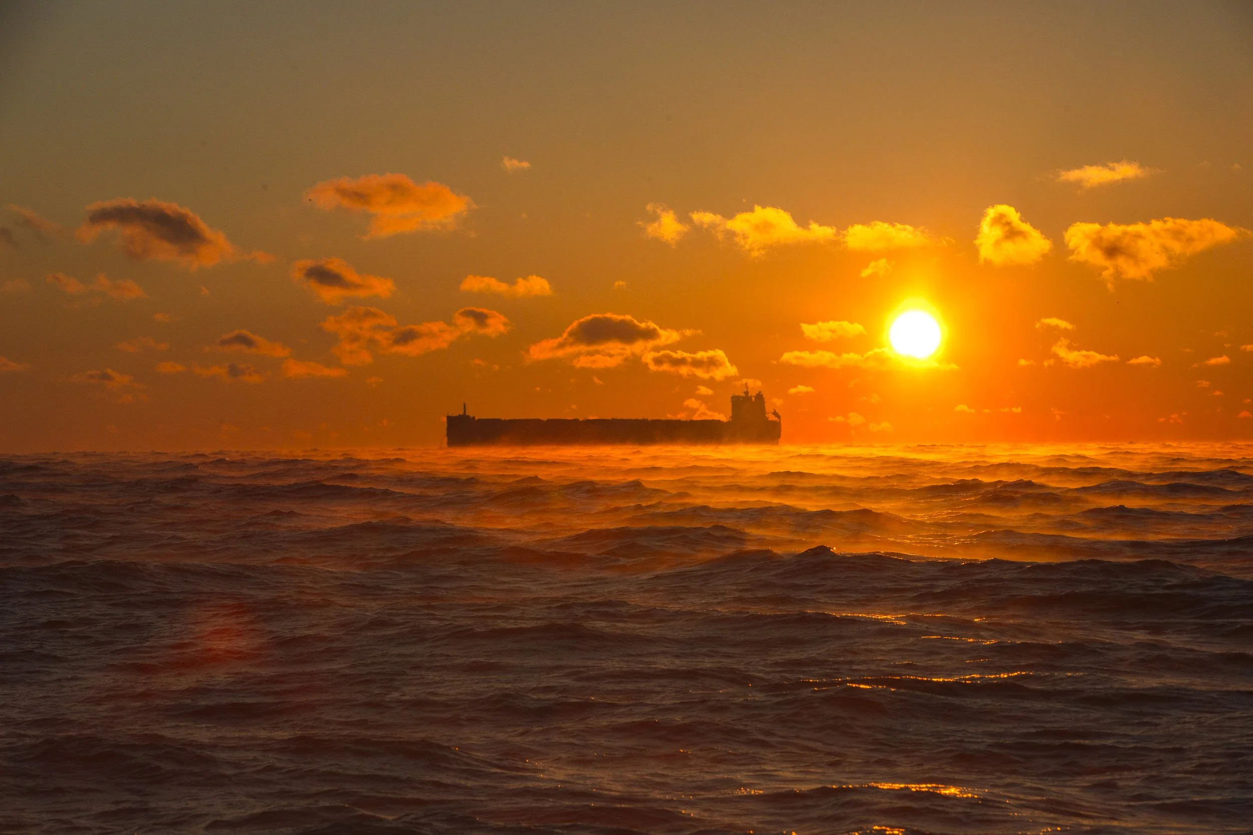 A ship sailing in the ocean during a vibrant sunset with the sun near the horizon, colorful sky with scattered clouds, and choppy waves.