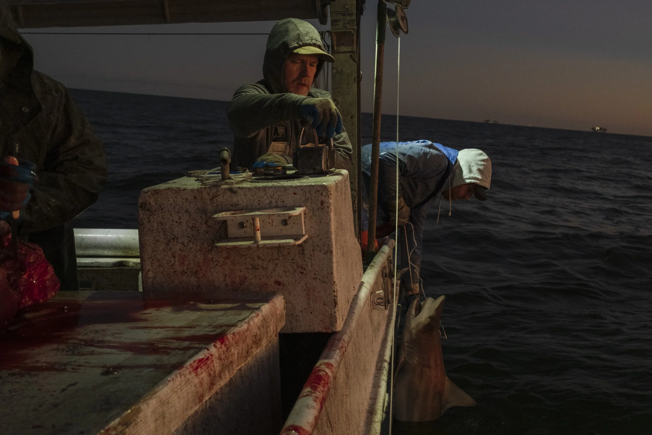 Two men on a boat at dusk, one is fishing with a rod hanging over the side, and the other is adjusting equipment on the boat.