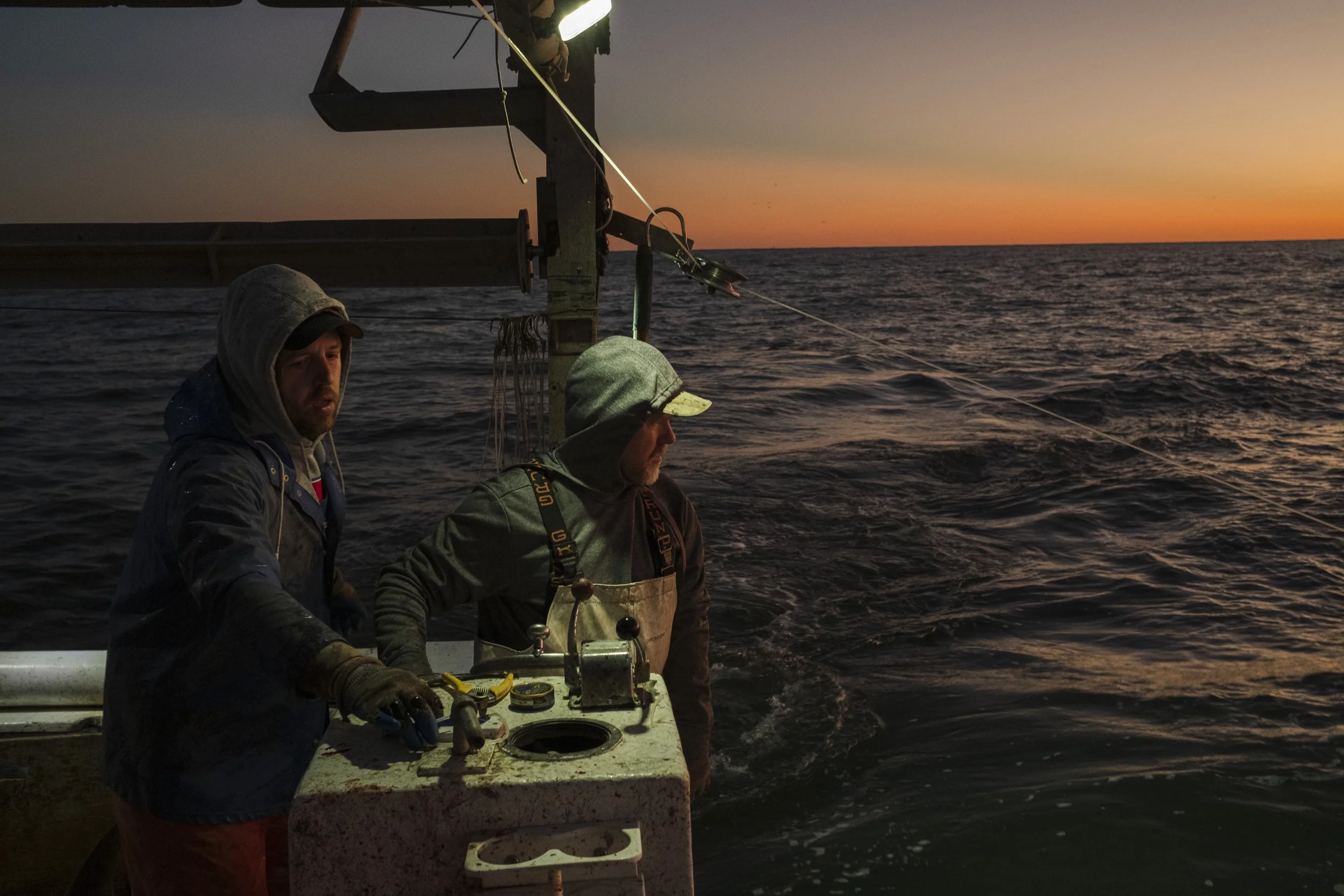 Two men on a boat at sunset, working with fishing equipment, with the ocean and colorful sky in the background.
