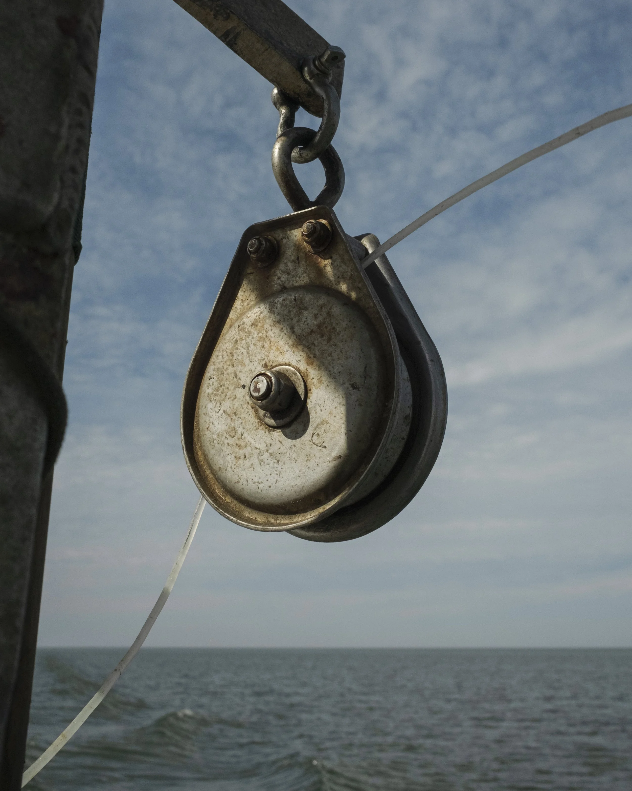 Close-up of a metal pulley and chain on a boat with water and cloudy sky in the background.