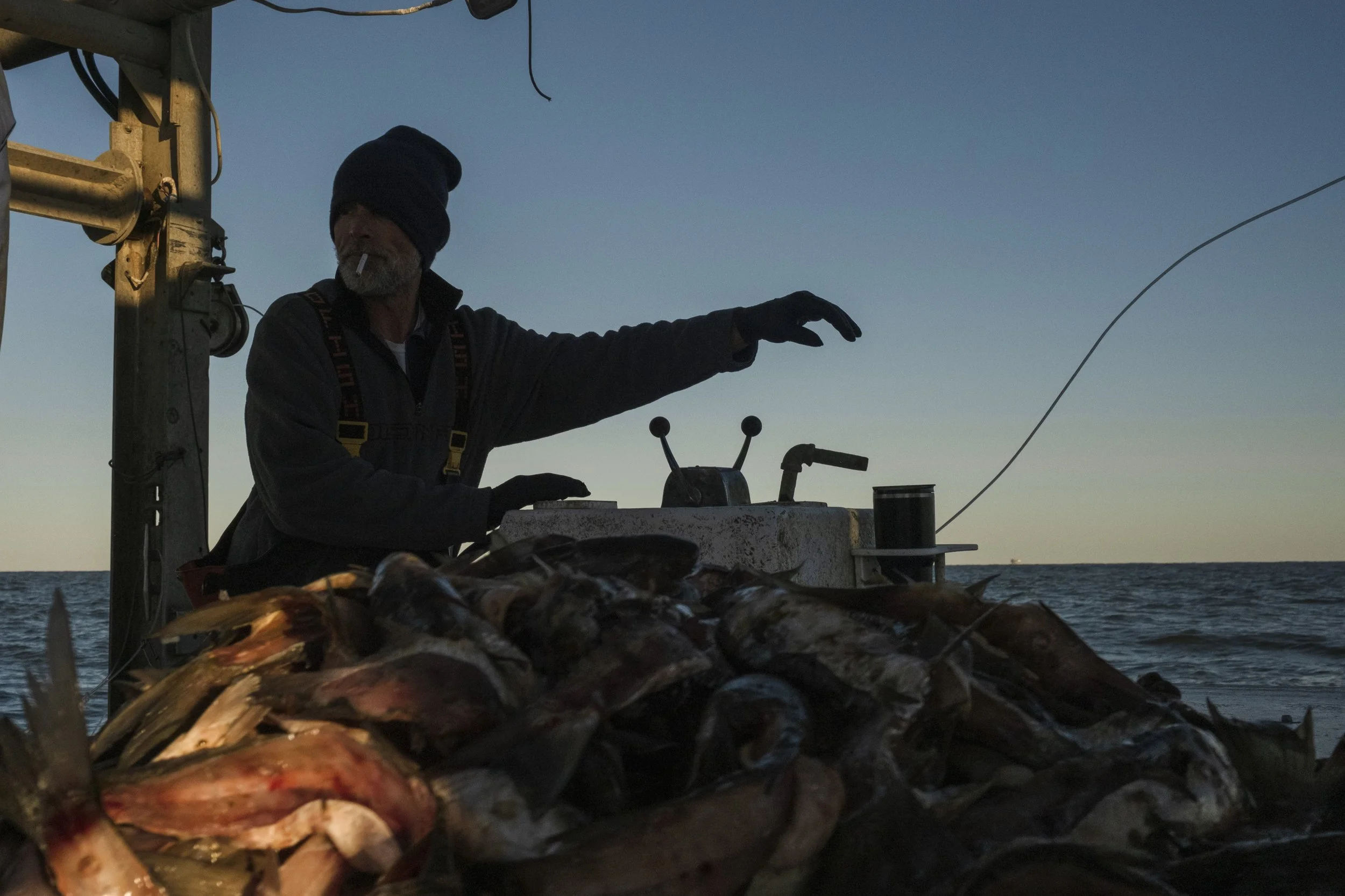 A man wearing a beanie and gloves on a boat, smoking a cigarette, surrounded by a pile of fish with the ocean and sky in the background at dusk or dawn.