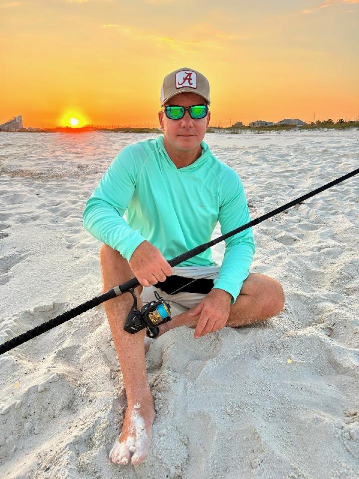 A man sitting in white sand on a beach during sunset, holding a fishing rod. He is wearing a gray cap with a red 'A' logo, reflective sunglasses, a light blue long-sleeve shirt, and shorts.