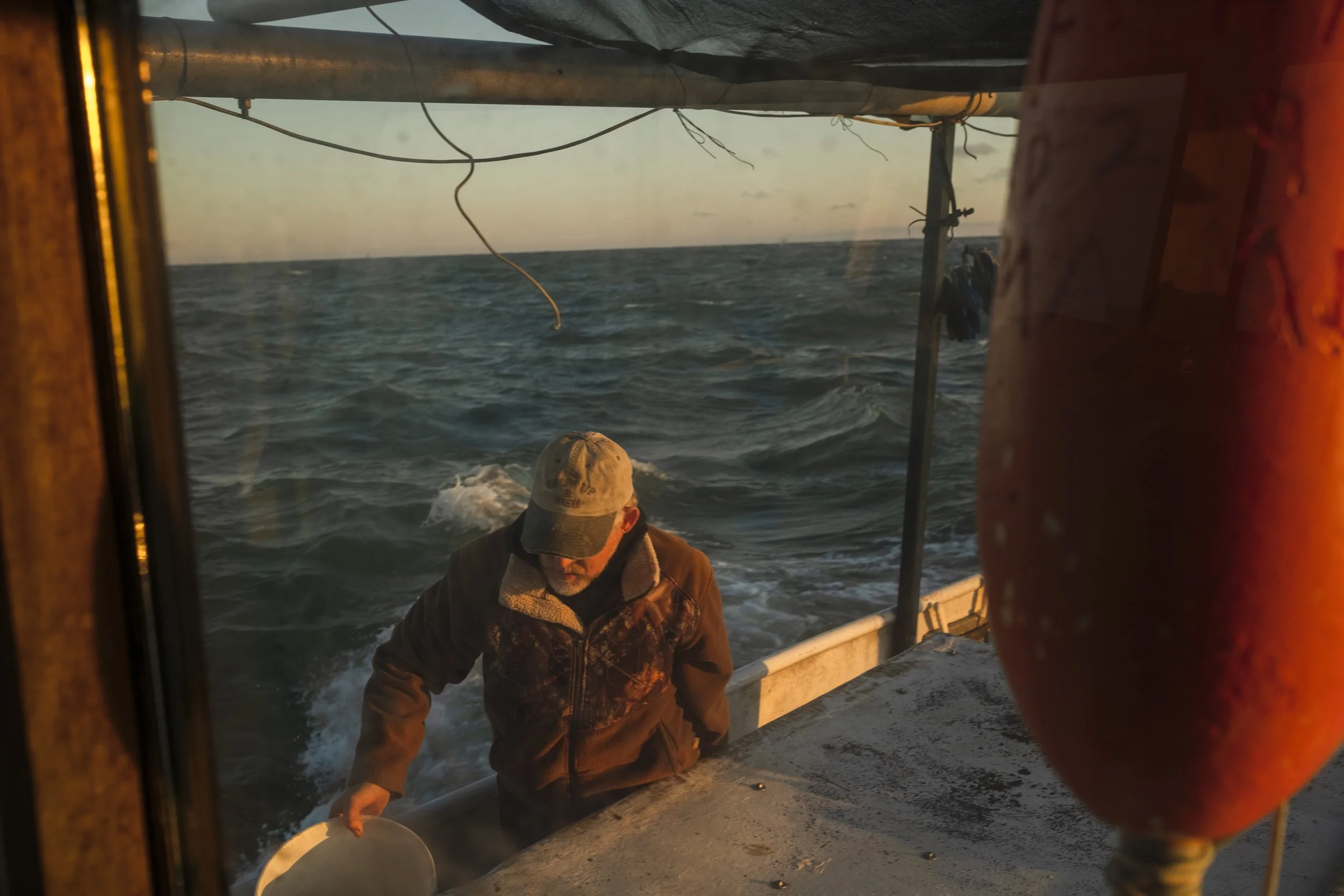 A man on a boat at sea during sunset, wearing a cap and jacket, holding a small bucket, with the ocean waves in the background.